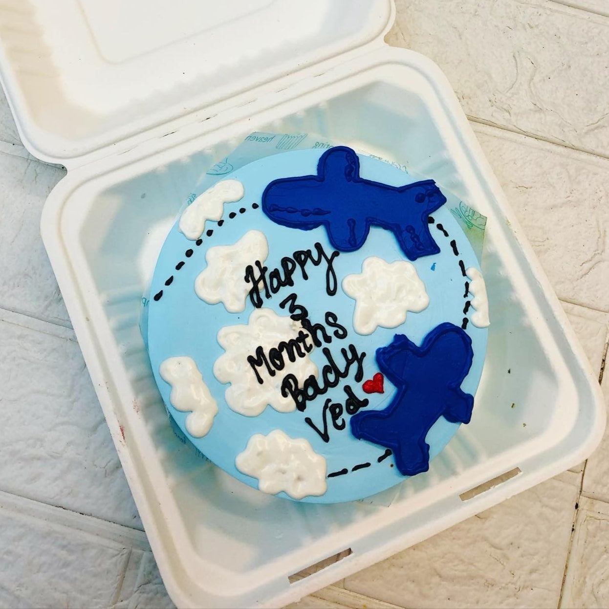 A blue-themed cake with airplane decorations and a message celebrating a 3-month milestone, placed inside a white bento box.