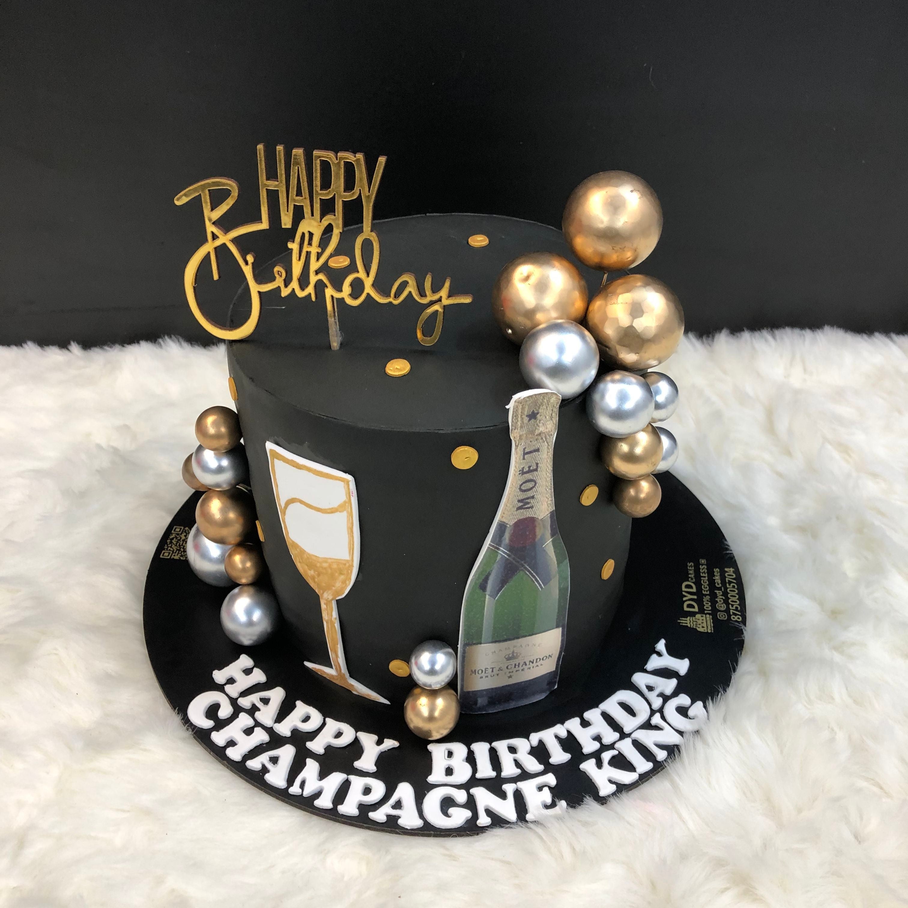 A black birthday cake decorated with gold and silver spheres, featuring a "Happy Birthday" topper, a champagne bottle, and a champagne glass design. The cake is placed on a black base with the text "Happy Birthday Champagne King."