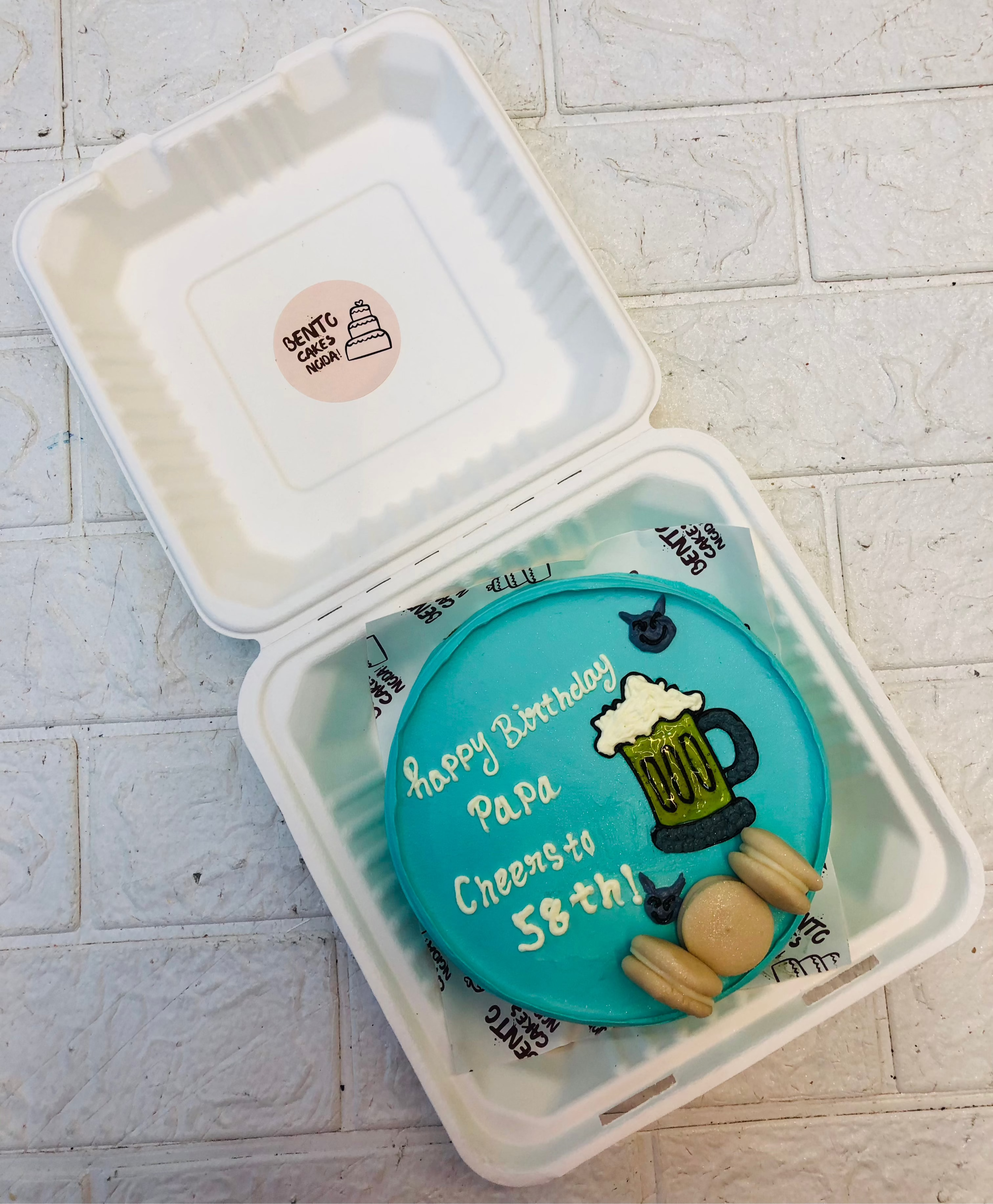 A festive blue birthday cake with a beer mug design, featuring the message 'Happy Birthday Papa Cheers to 58th!' inside a takeout box from Bentocake, garnished with macarons and a tiny bear figure."
This title and alt text highlight the fun and celebratory aspects of the cake while providing a clear description. If you’d like any tweaks or a different style, just let me know!