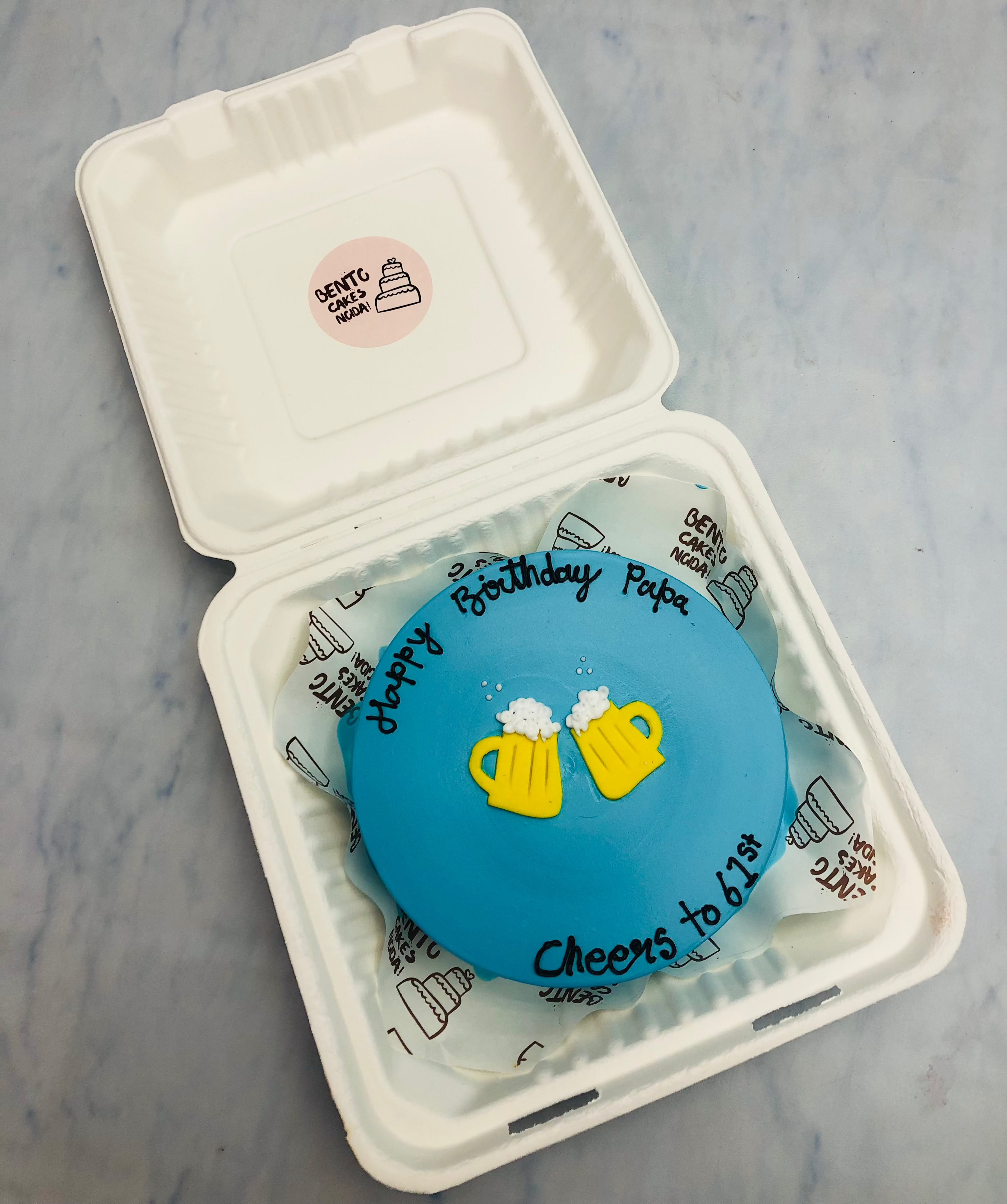 A vibrant blue birthday cake featuring the message 'Happy Birthday Papa' and 'Cheers to 61st' in black icing. The design includes two playful yellow beer mugs with foamy tops, adding a fun celebratory touch.