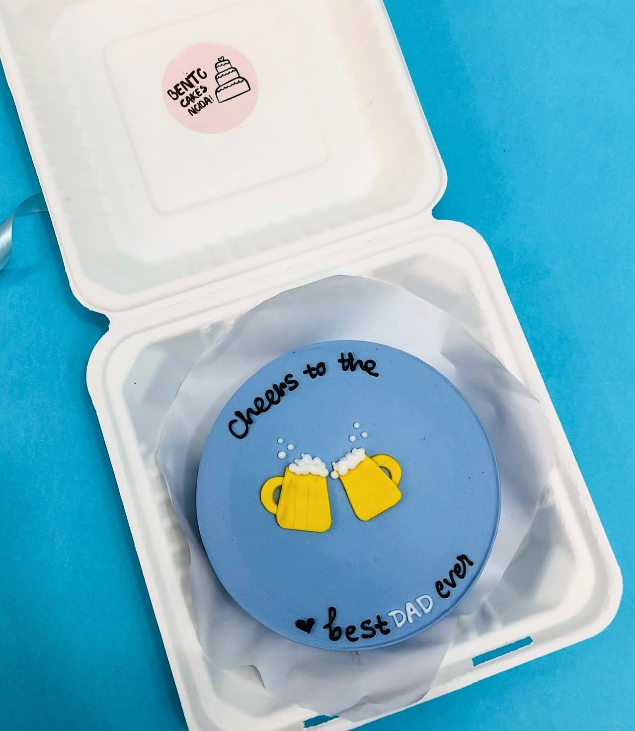A round cake with a blue icing top, featuring two yellow beer mugs clinking together. The cake has the message "Cheers to the best DAD ever" written in black and blue icing. 