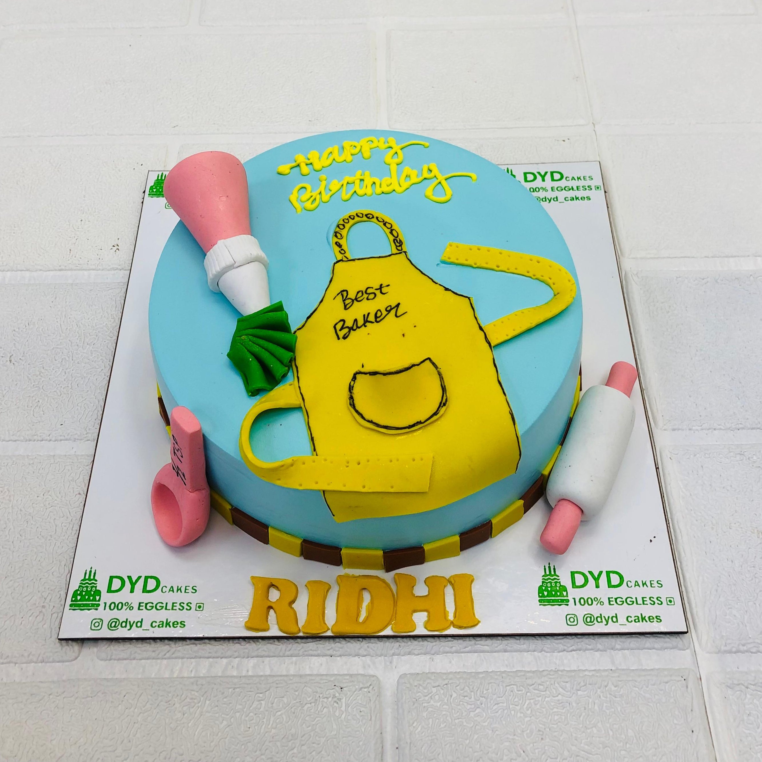 A sky-blue color cake is designed with kitchen utensils on its top and surround having an apron on its top and a text "RIDHI" written on the side of cake.
