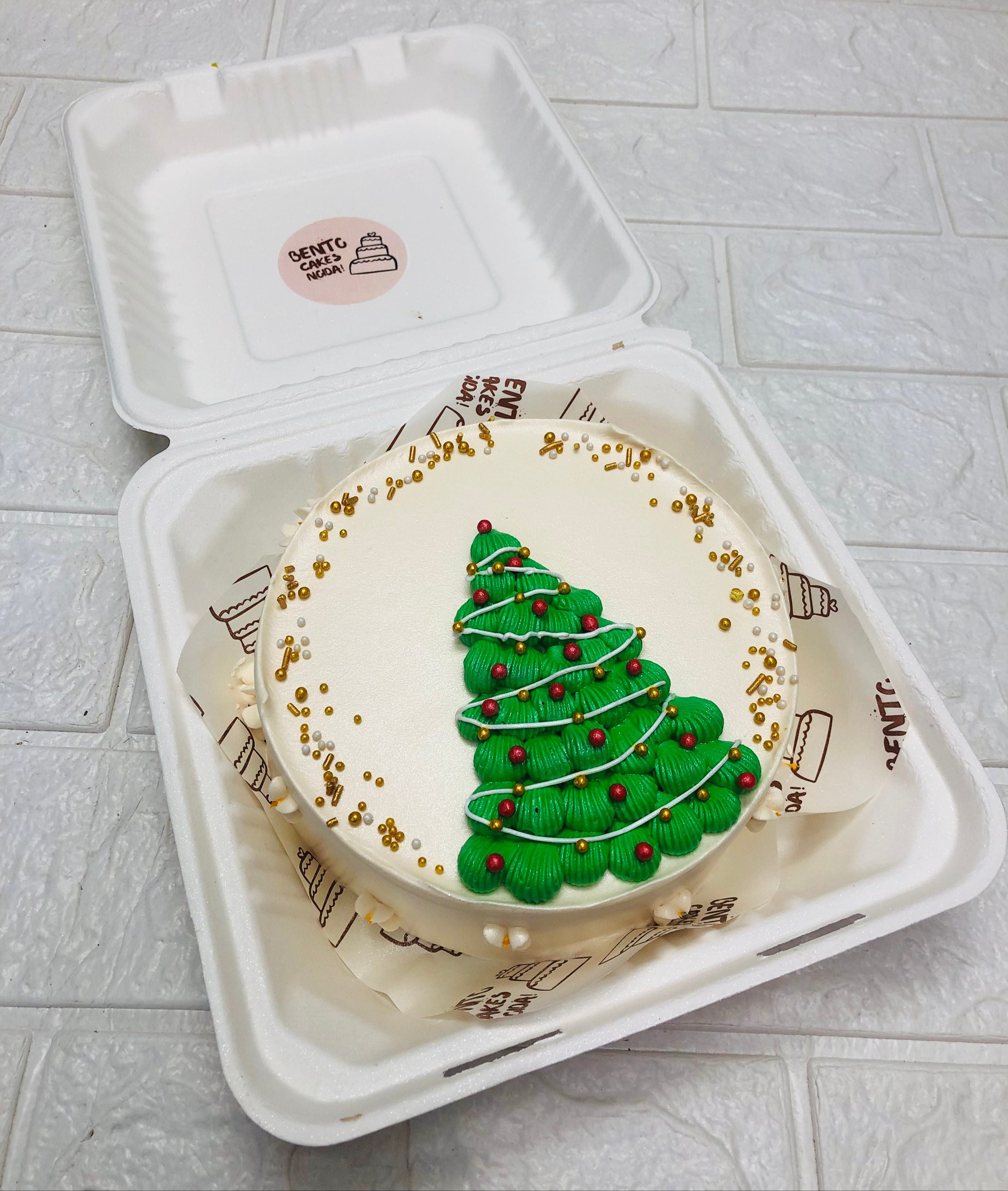 An offwhite bento cake is designed on christmas tree theme having a green tree design on cake and decorated with golden and red sprinkles.
