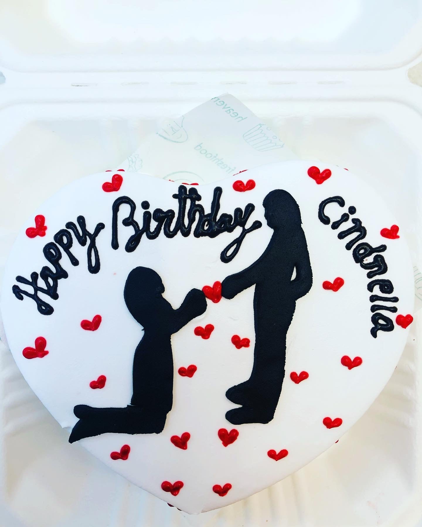 A white heart shape cake is decorated with red hearts and a design of 2 persons made with fondant on it.