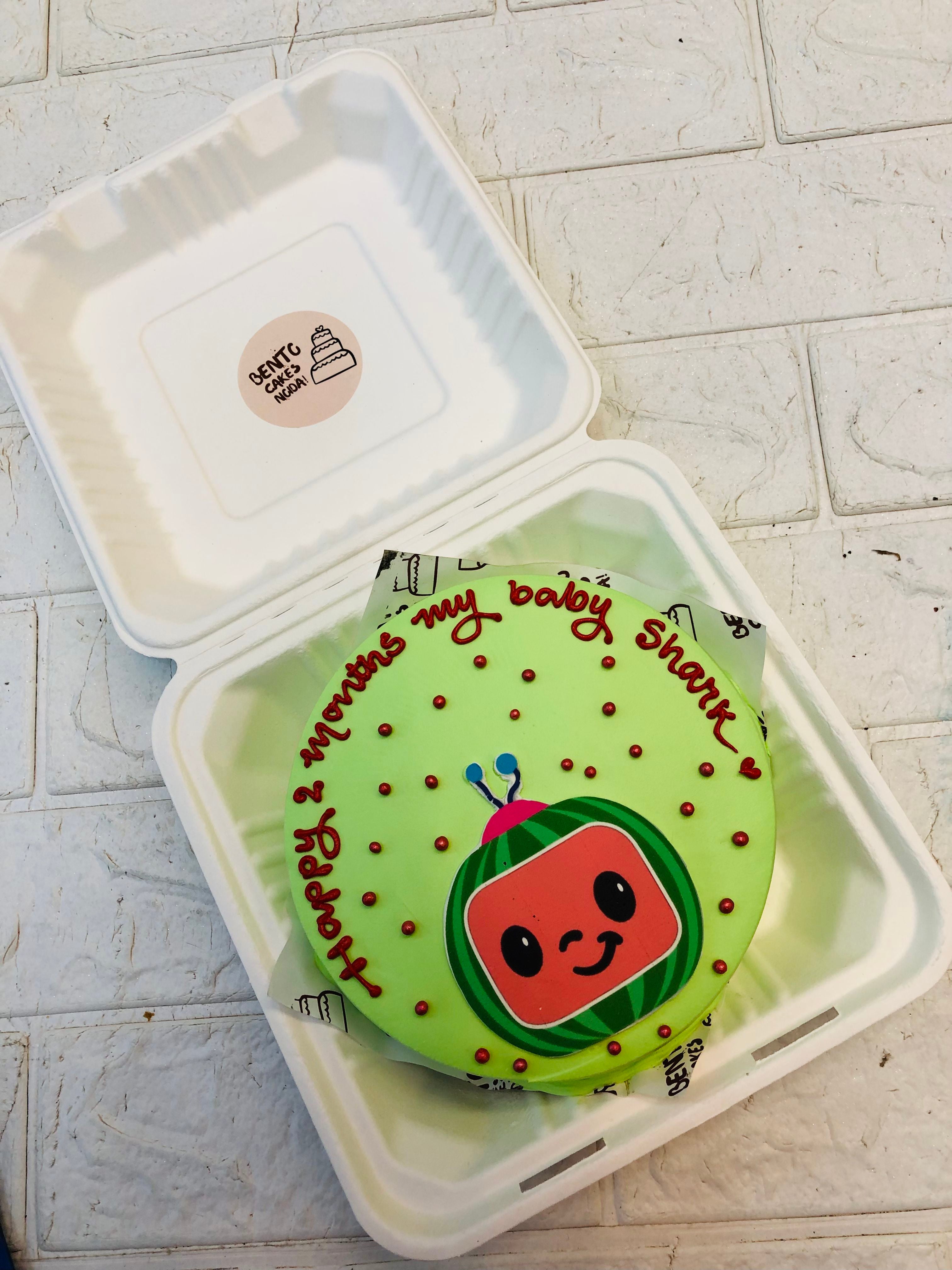 A light green bento cake with a cocomelon print and deecorated with red sprinkles.