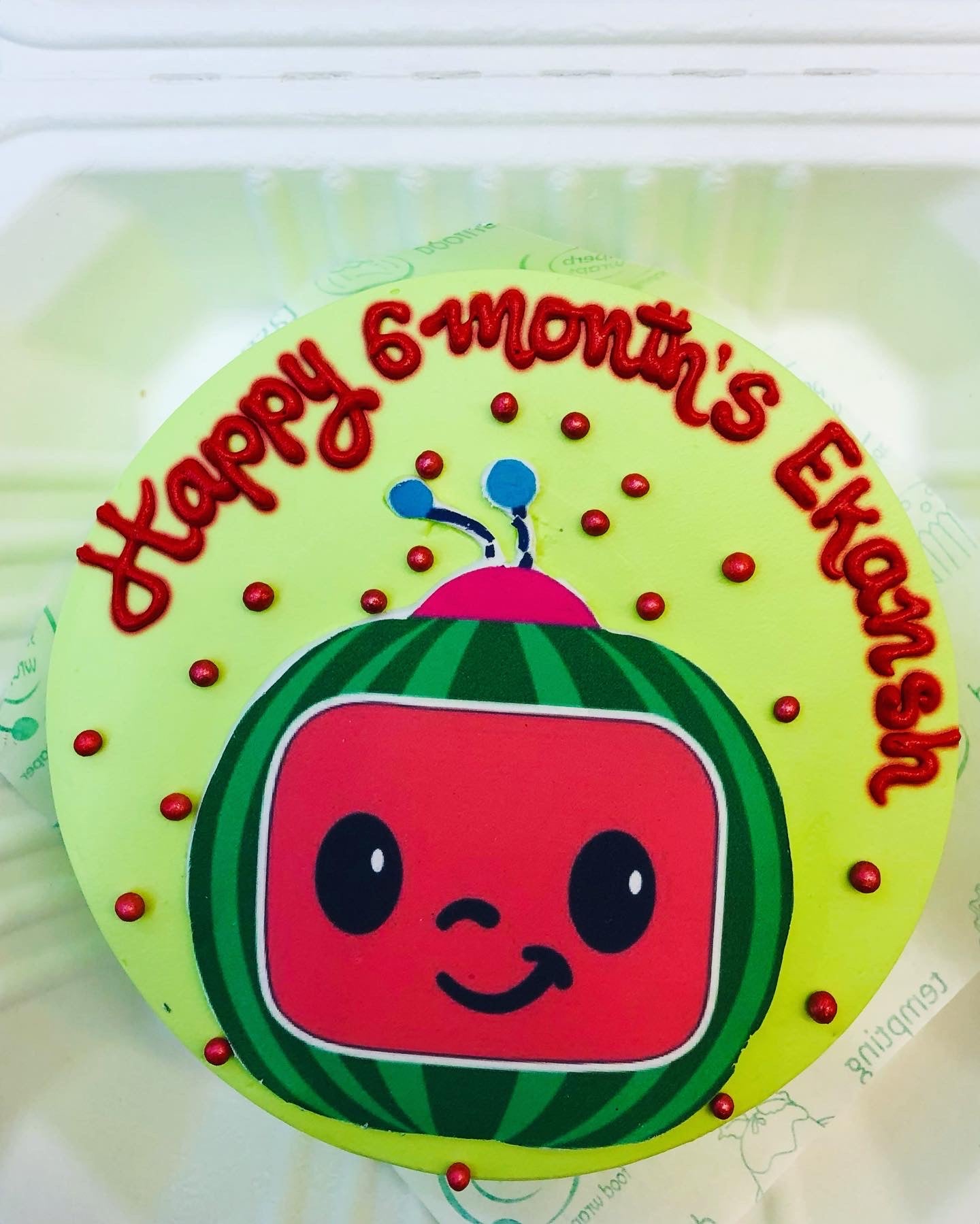 A light green cake is decorated with a Coco melon cartoon sticker placed on it and a Text of Happy 6 Months Eksnsh written with red icing on it.