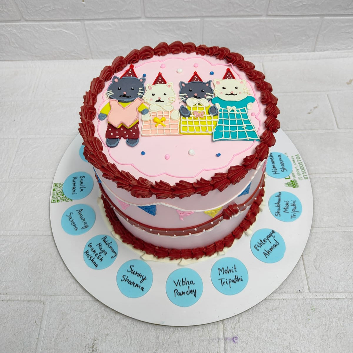 A beautifully decorated birthday cake with a pink top layer featuring four cartoon cats wearing party hats and vibrant outfits. The cake is bordered with red icing, with personalized names inscribed on blue circles around the base.