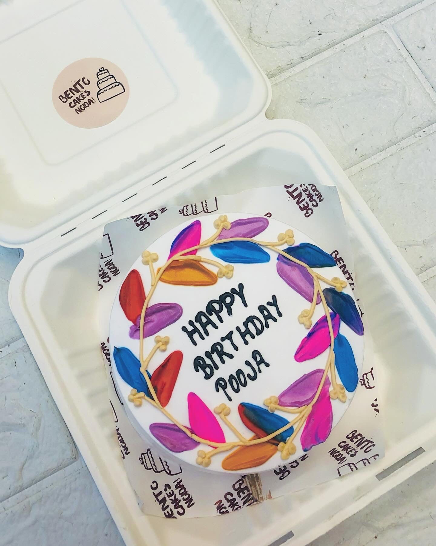 A birthday cake with the message 'Happy Birthday Pooja' written on it, surrounded by colorful leaf decorations in shades of pink, purple, blue, orange, and red.