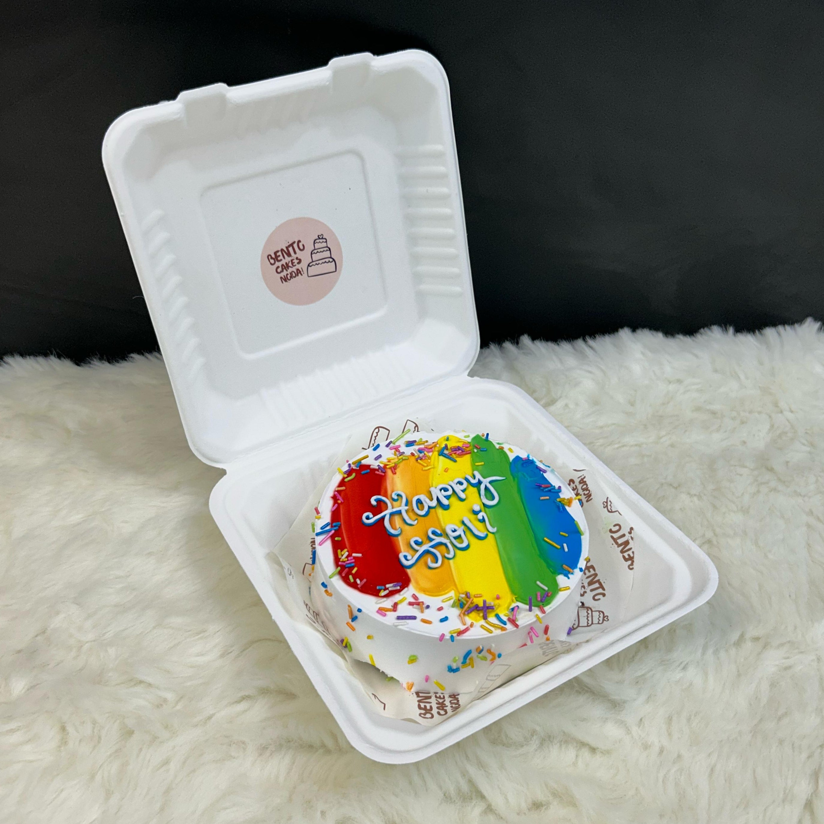 A colorful bento cake in a white box with the text "Happy Holi" written on it. The cake features vibrant rainbow stripes and is topped with multicolored sprinkles.