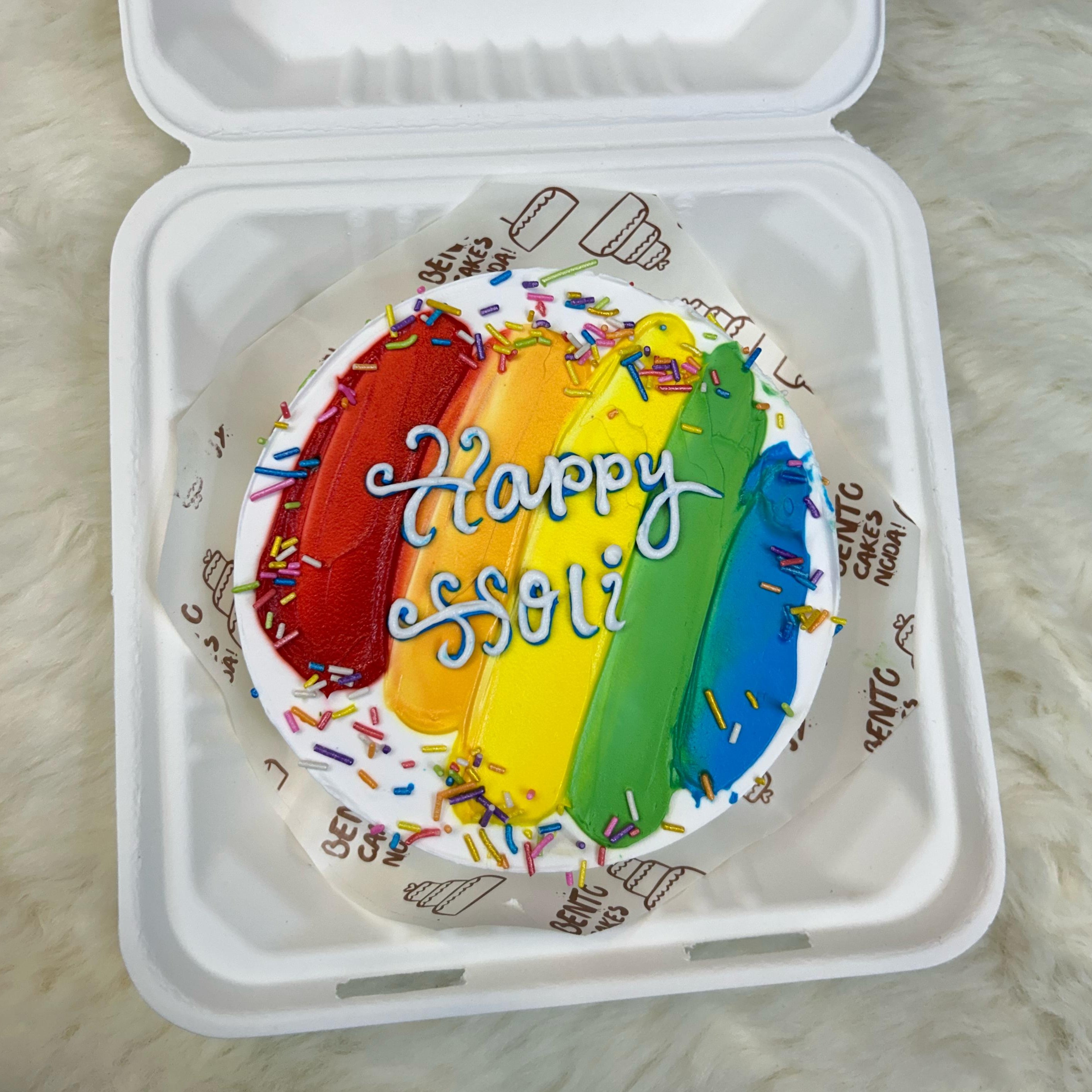 A colorful bento cake in a white box with the text "Happy Holi" written on it. The cake features vibrant rainbow stripes and is topped with multicolored sprinkles with front view.