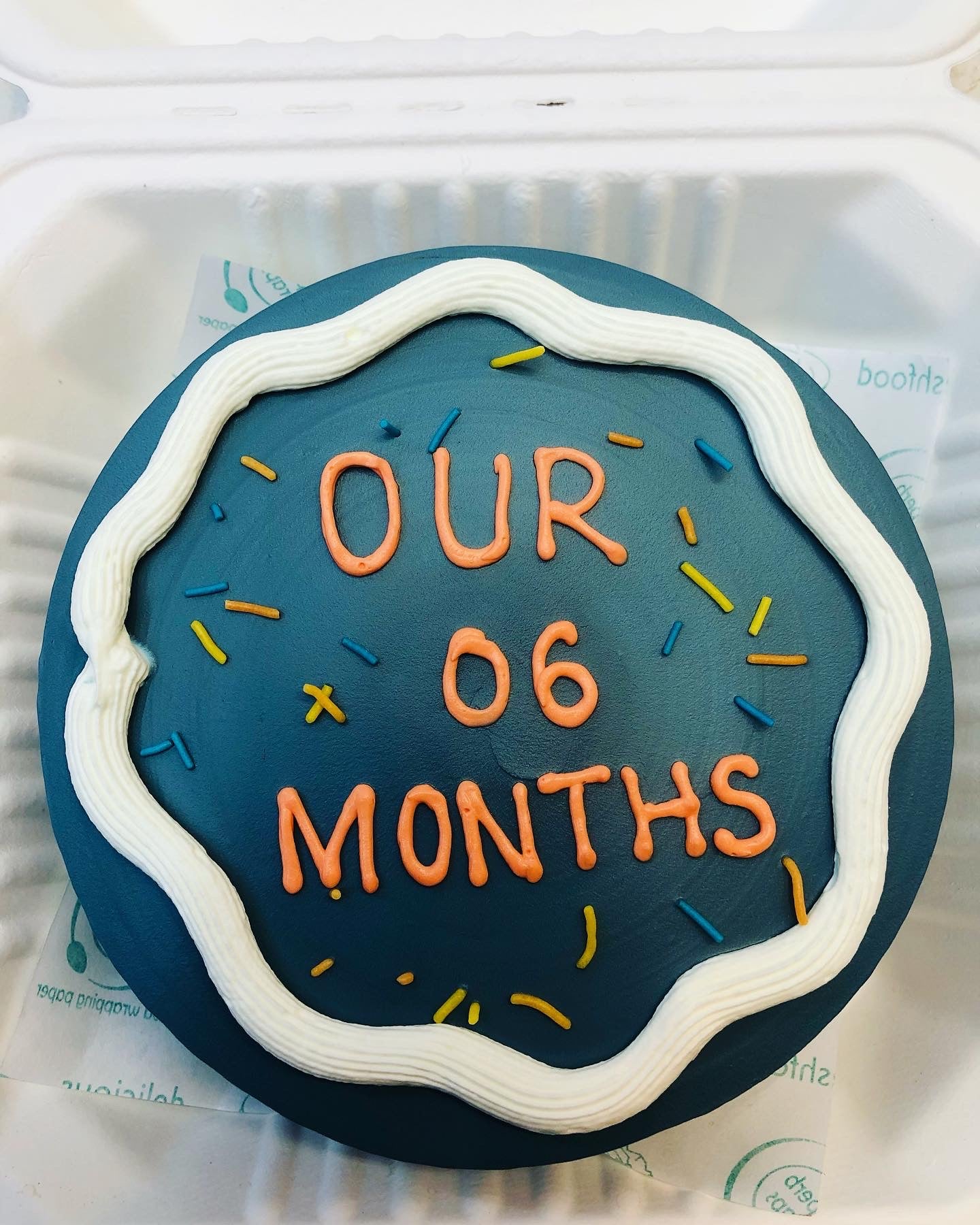A dark blue cake in a bento box decorated perfectly and a text "OUR 06 MONTHS" beautifully written on it.