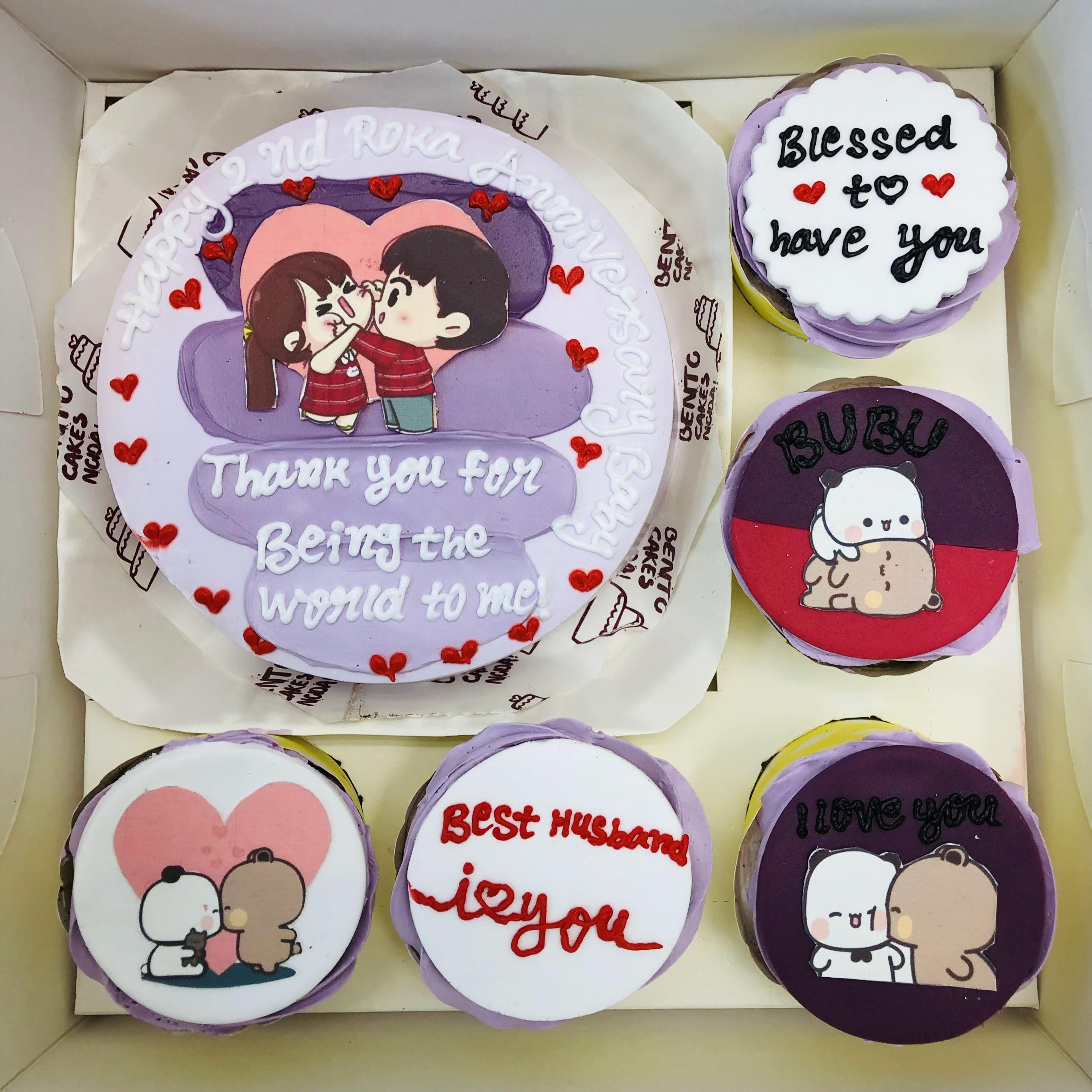 A romantic bento cake set featuring one large cake and four smaller cupcakes, decorated with love-themed designs. The large cake displays an illustration of a couple wearing red shirts, surrounded by the text "Happy 2nd Roka Anniversary" and the heartfelt message "Thank you for being the world to me!" The smaller cupcakes include unique messages such as "Blessed to have you," "Best Husband," and "I love you," alongside adorable illustrations of hugging characters and playful designs.