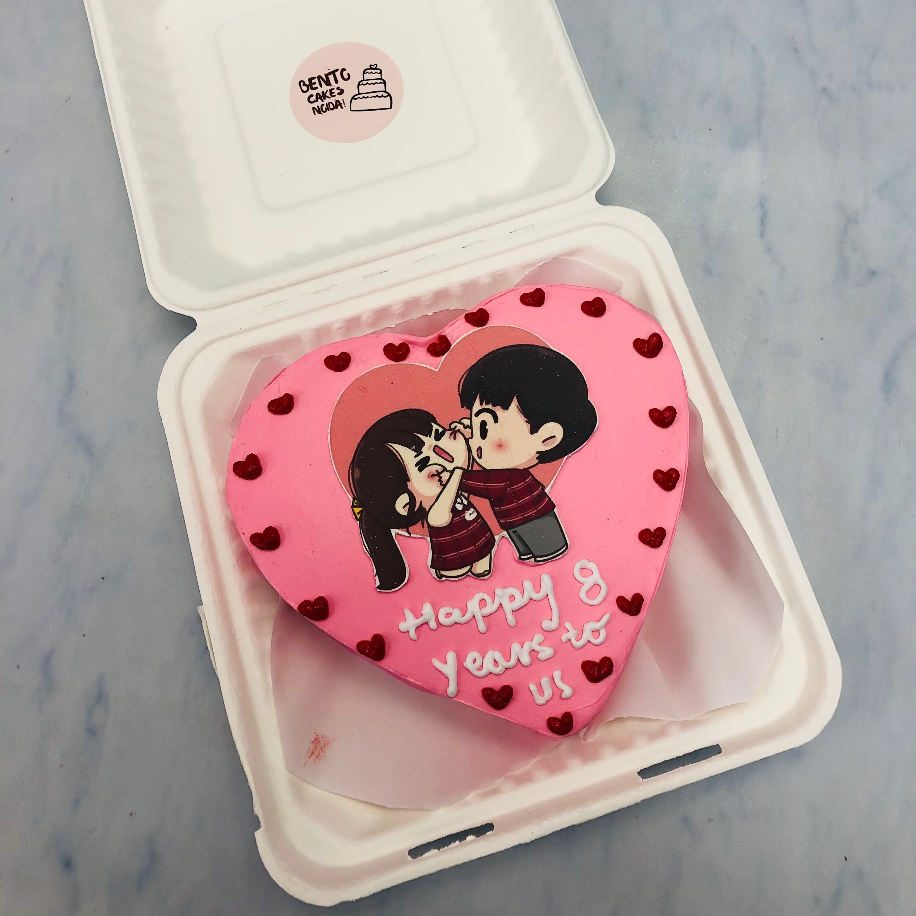 A heart shape cake in pink color having cute couple sticker and is decorated with little red hearts on it's boundary.