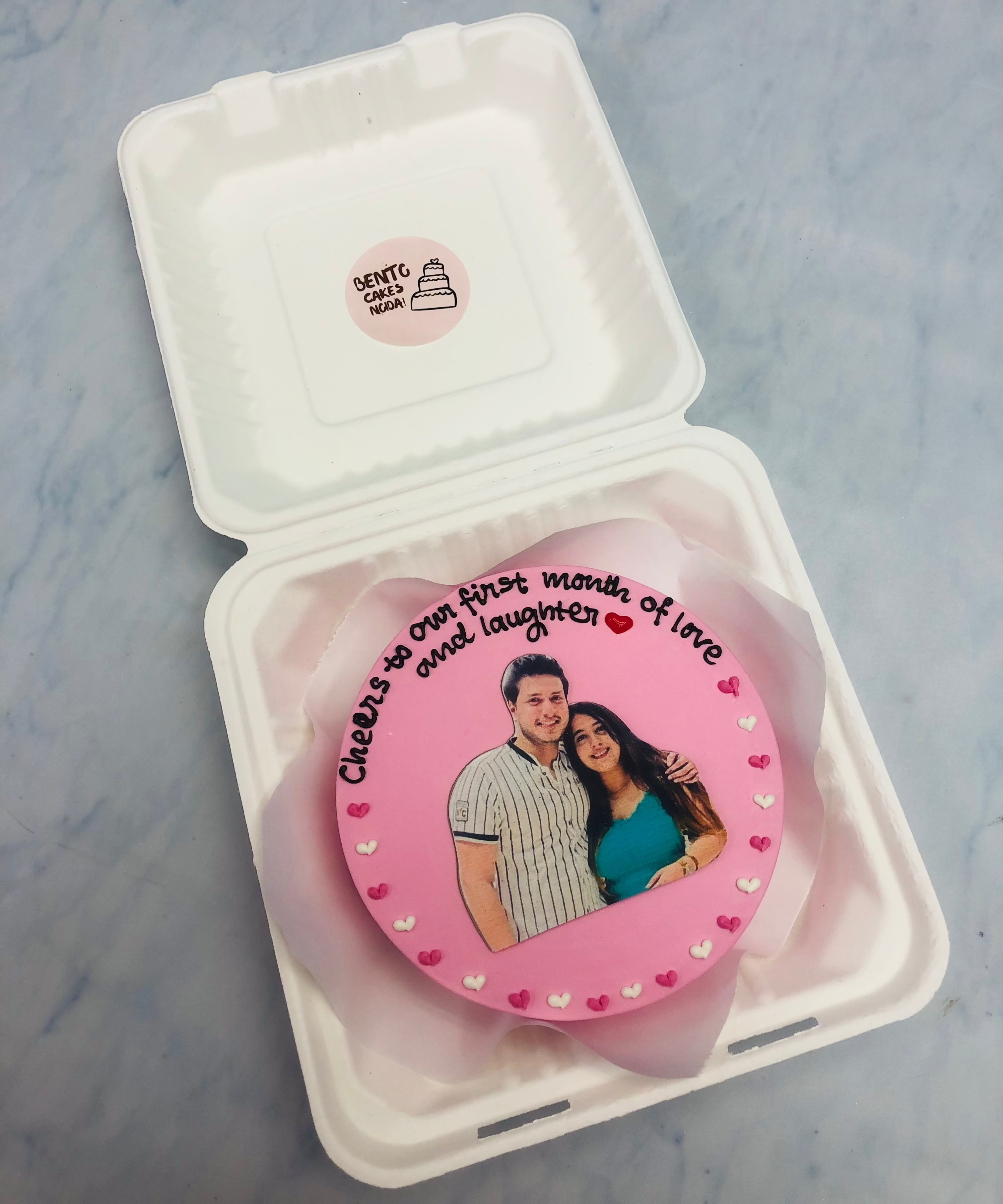 A charming pink bento cake inside a white box, featuring a printed image of a couple, surrounded by delicate heart decorations, with the message "Cheers to our first month of love and laughter."