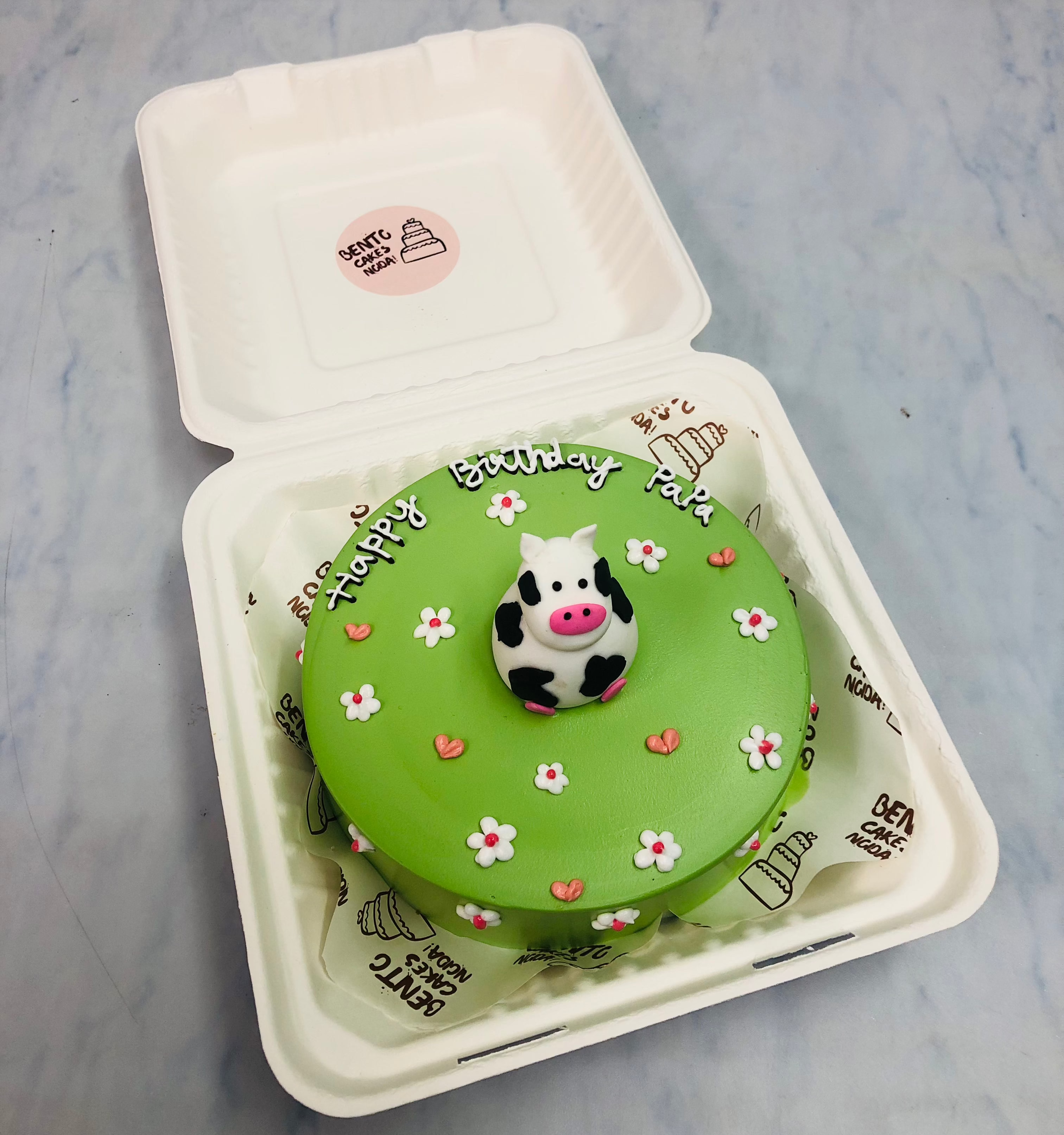 A green birthday cake with a cow figurine on top, decorated with small white flowers and pink hearts, placed inside a bento box. The cake has the text "Happy Birthday Papa" written on it.