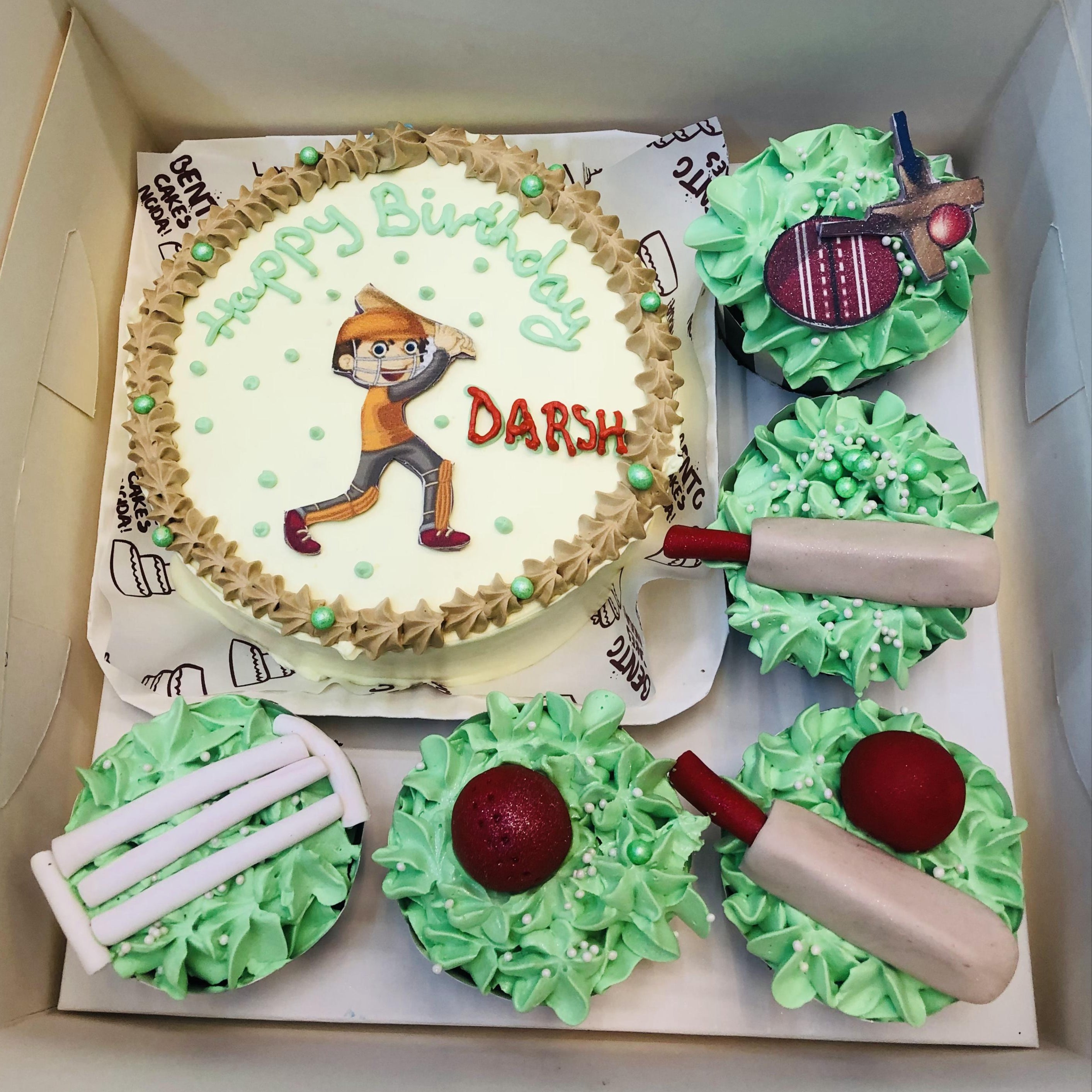 A cricket-themed bento cake set featuring a round cake with a white base, green dots, and a brown border, displaying an illustration of a cricket player. The cake has the text "Happy Birthday DARSH." Surrounding the main cake are six cupcakes with green frosting, decorated with cricket-related items like a bat, ball, and stumps.