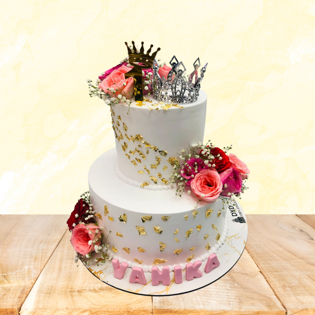 A two-tier white cake decorated with gold leaf accents, fresh pink and red roses, and baby's breath flowers. The top tier is adorned with a silver tiara and a gold crown. The base of the cake has the name "Vamika" written in pink letters. The cake is labeled as 100% eggless.