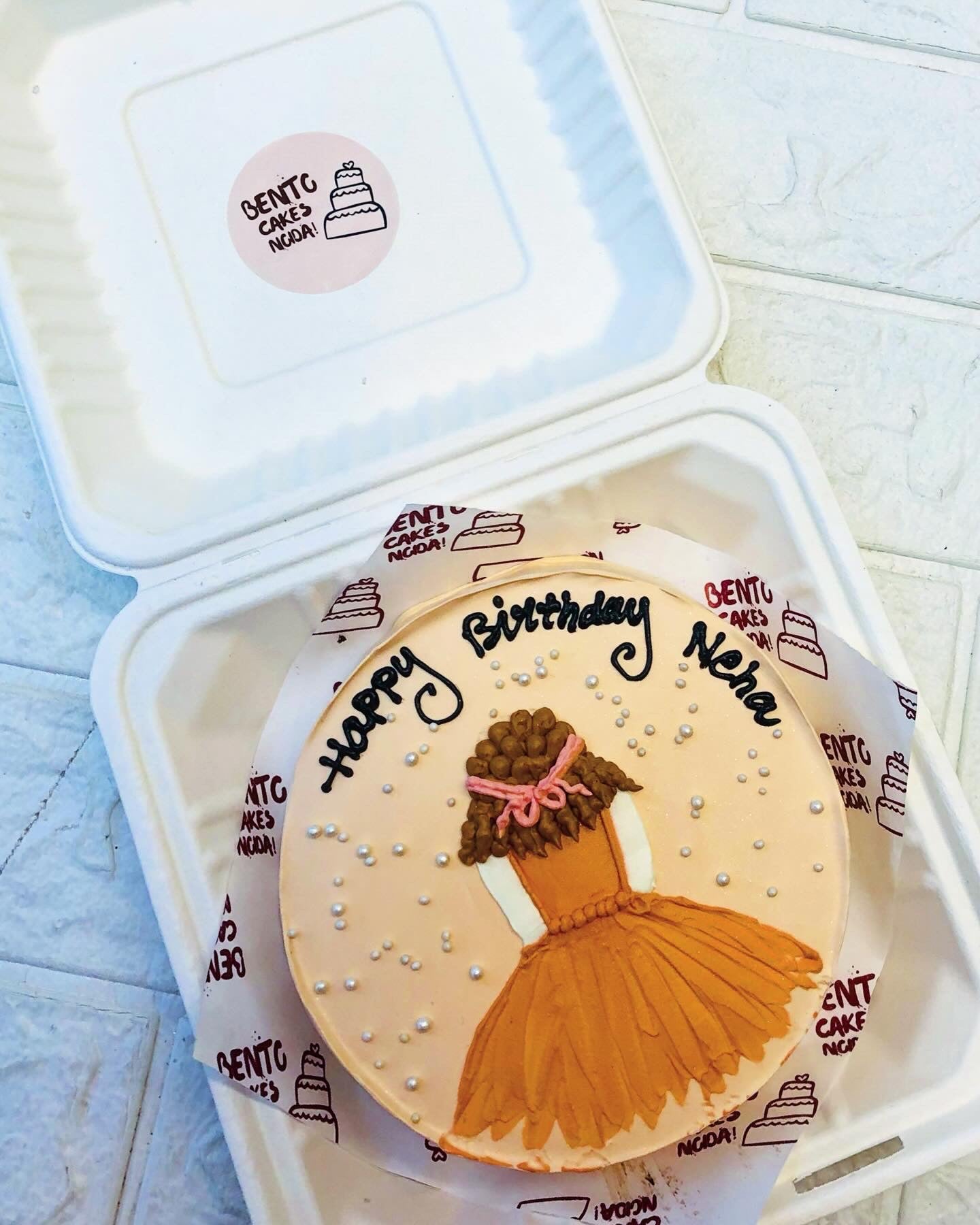 A custom birthday cake from Bento Cakes India, featuring an elegant design of a girl in a brown dress with curly hair and a pink bow. The cake is decorated with pearl-like dots and the message "Happy Birthday Nisha" written in black icing.