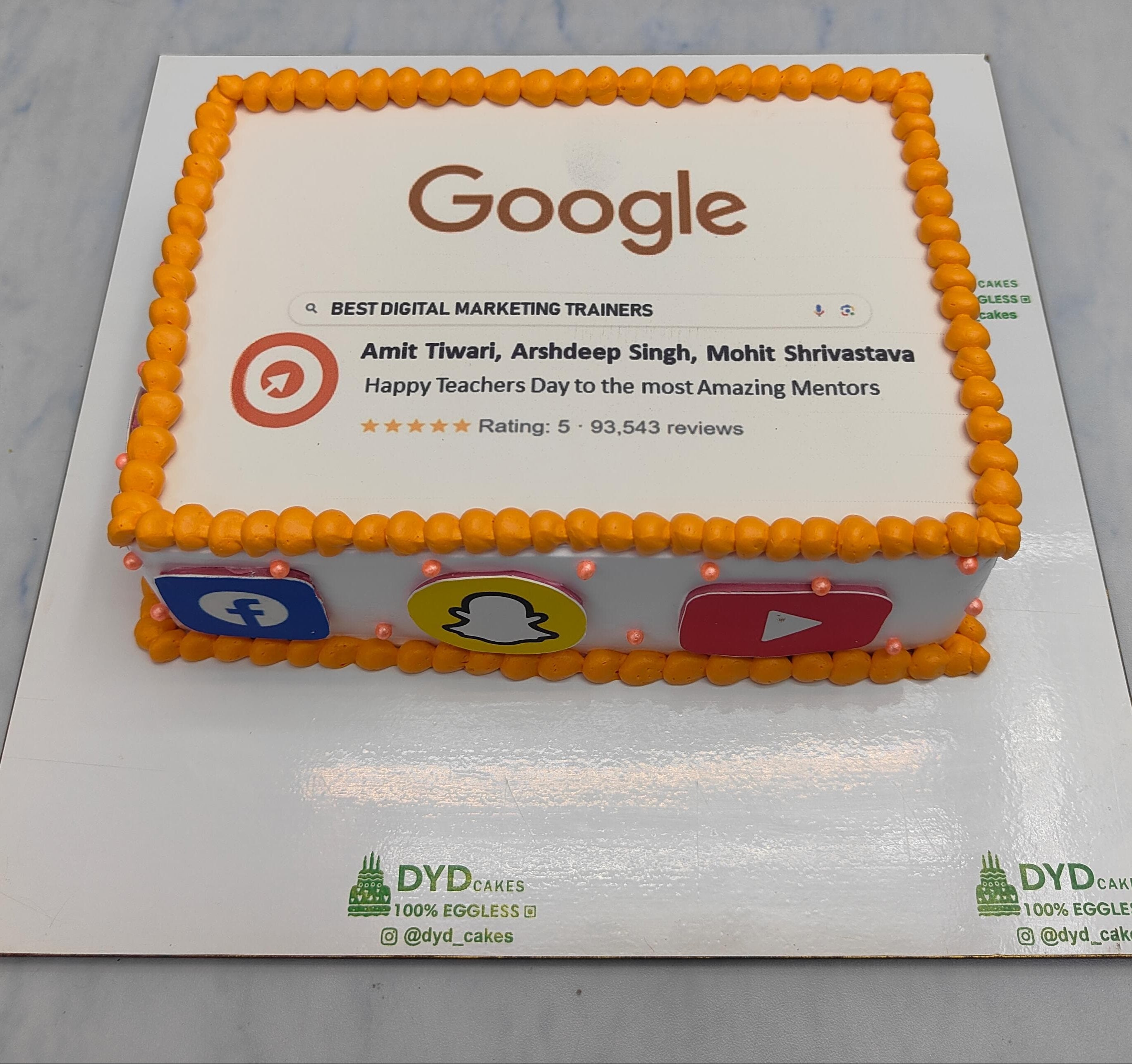 A rectangular cake decorated with a Google search result featuring Amit Tiwari, Arshdeep Singh, and Mohit Shrivastava as the best digital marketing trainers. The cake is adorned with orange icing and icons of Facebook, Snapchat, and YouTube on the sides.