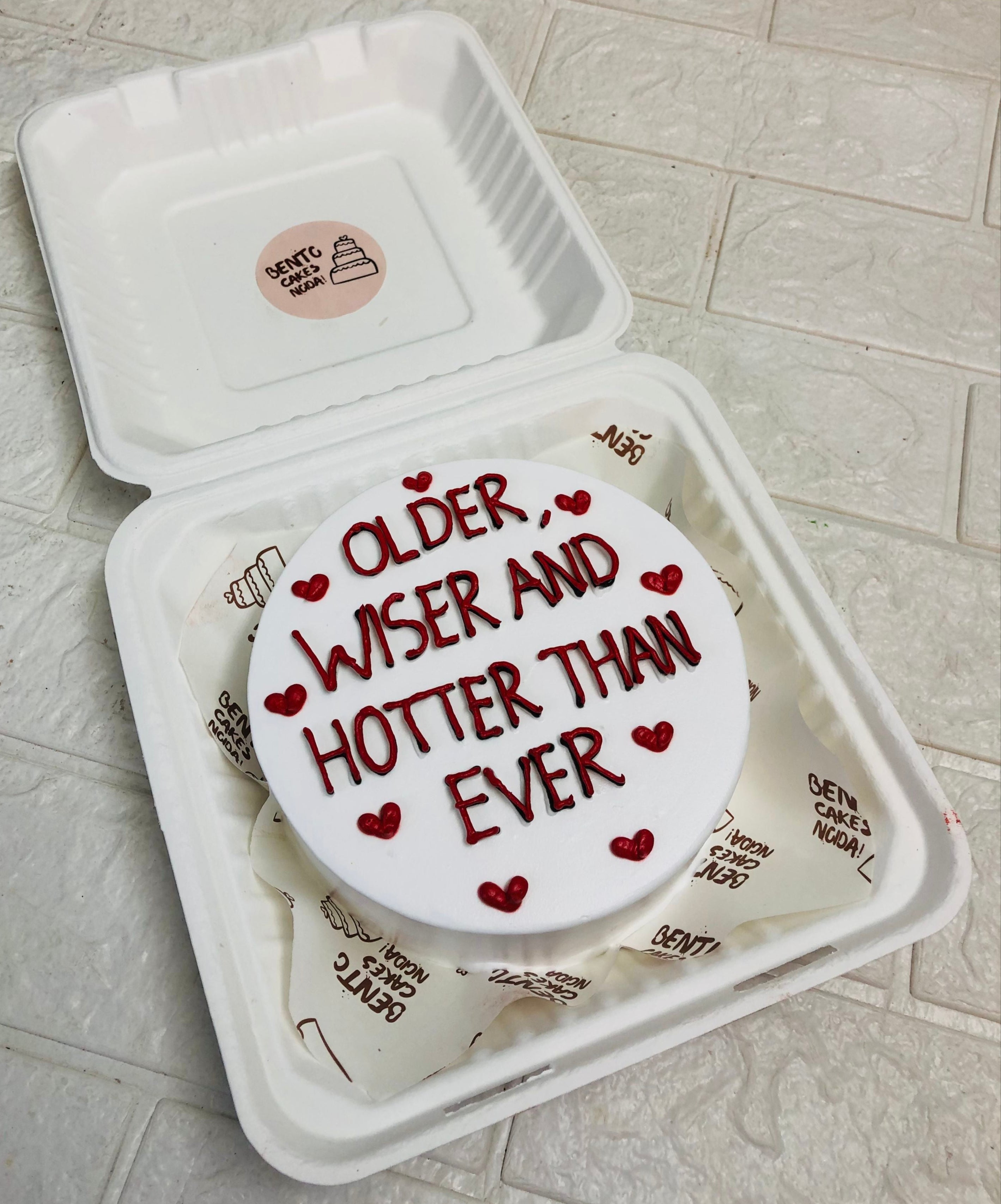 A white bento cake decorated with red hearts and a text "OLDER WISER AND HOTTER THAN EVER" written with red icing.