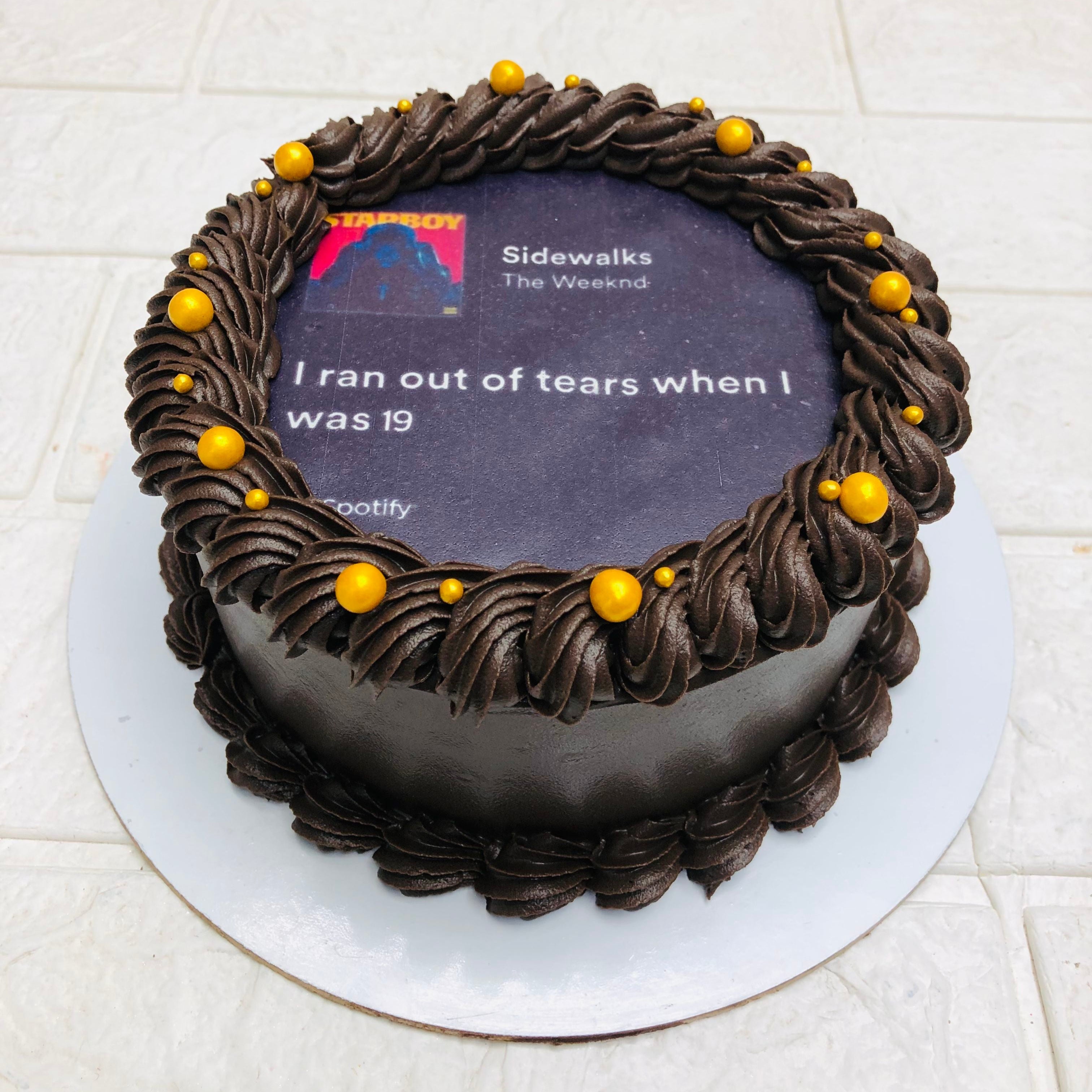 A round chocolate Fire cake with dark chocolate frosting and yellow decorative balls. The top of the cake features an edible image of a Spotify song playing, showing "Sidewalks" by The Weeknd, with the text "I ran out of tears when I was 19."