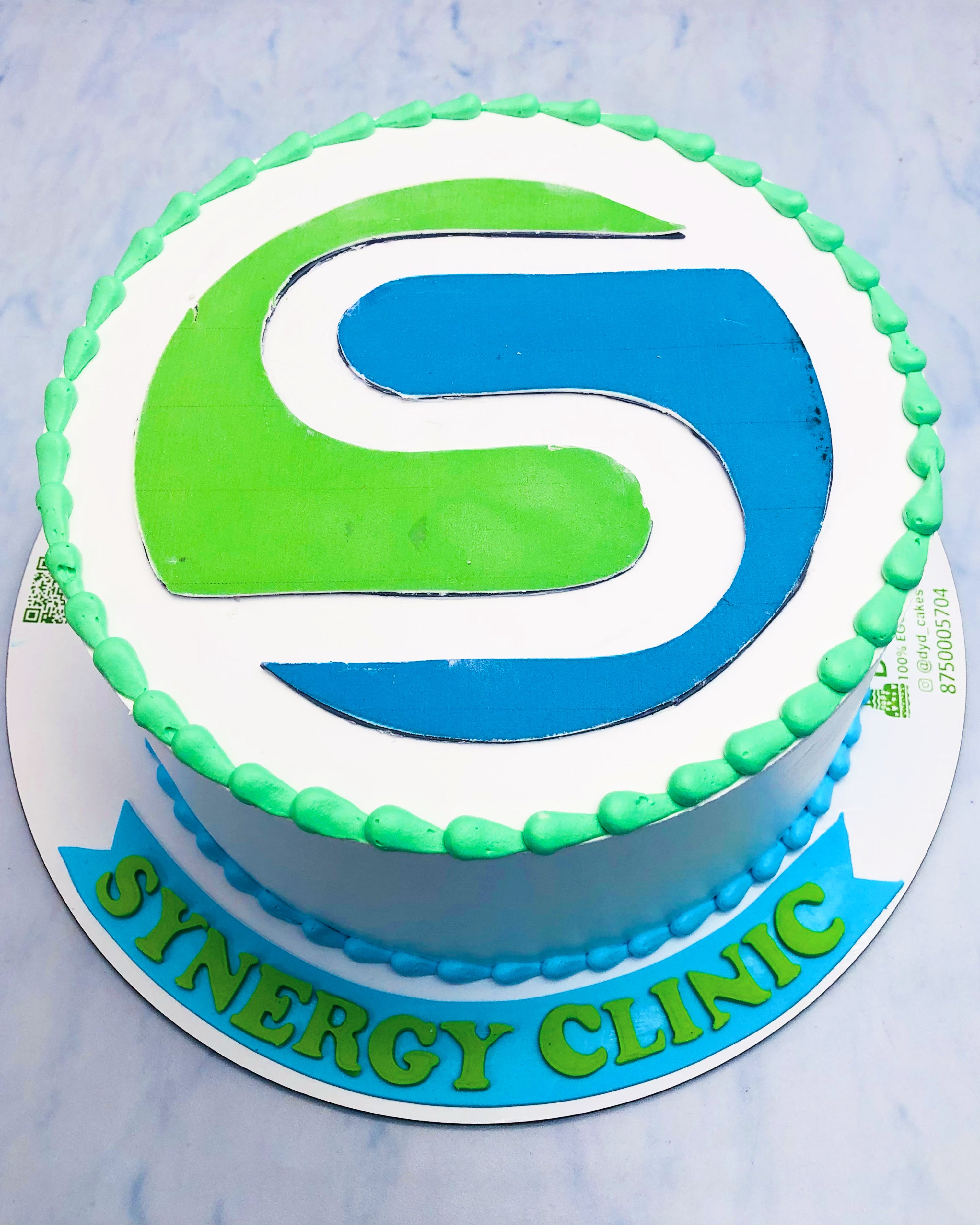 A round cake with the Synergy Clinic logo in green and blue icing on top, bordered with green icing dots, and the words 'SYNERGY CLINIC' written in green on a blue ribbon at the base.