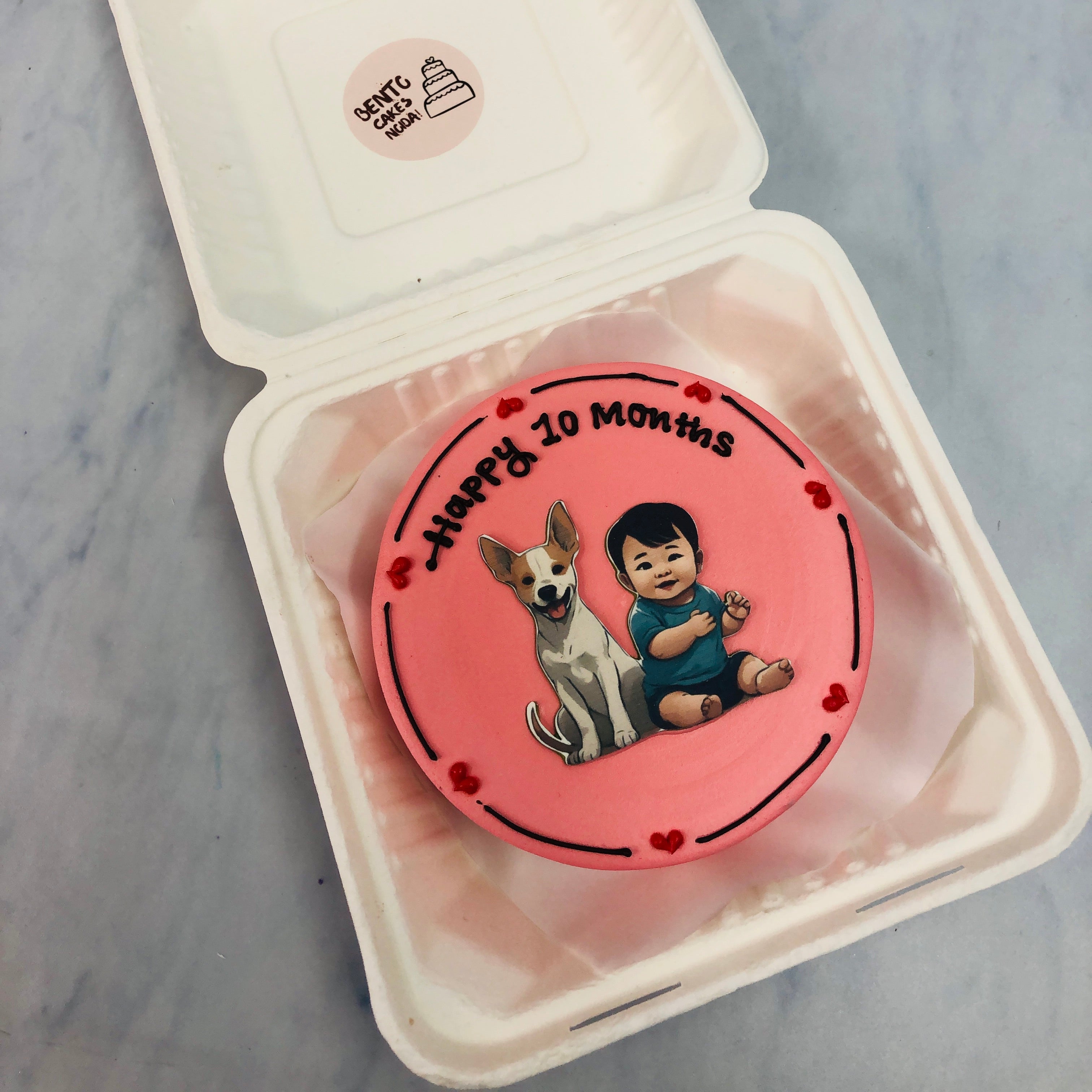 A light red color cake with a baby and a dog print on it, designed with red hearts on it's boundary.