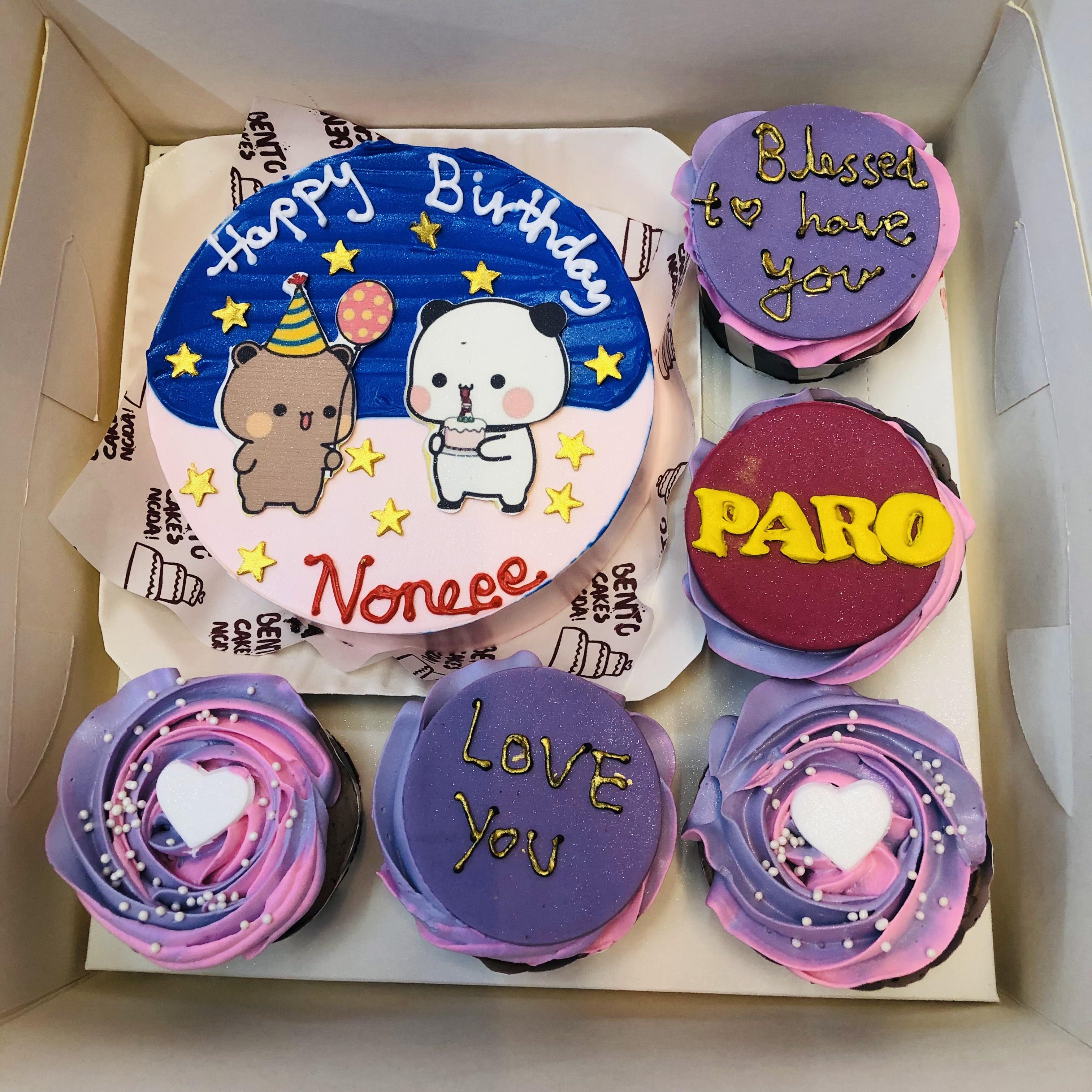 A bento cake set featuring a colorful birthday cake with two cartoon characters—a bear and a panda wearing party hats—on a pink and blue frosted base. The text reads "Happy Birthday Noneee." Accompanying the cake are five cupcakes, each uniquely designed with messages like "Blessed to have you," "PARO," and "LOVE YOU," along with decorative frosting in purple, pink, and white with heart-shaped sprinkles.