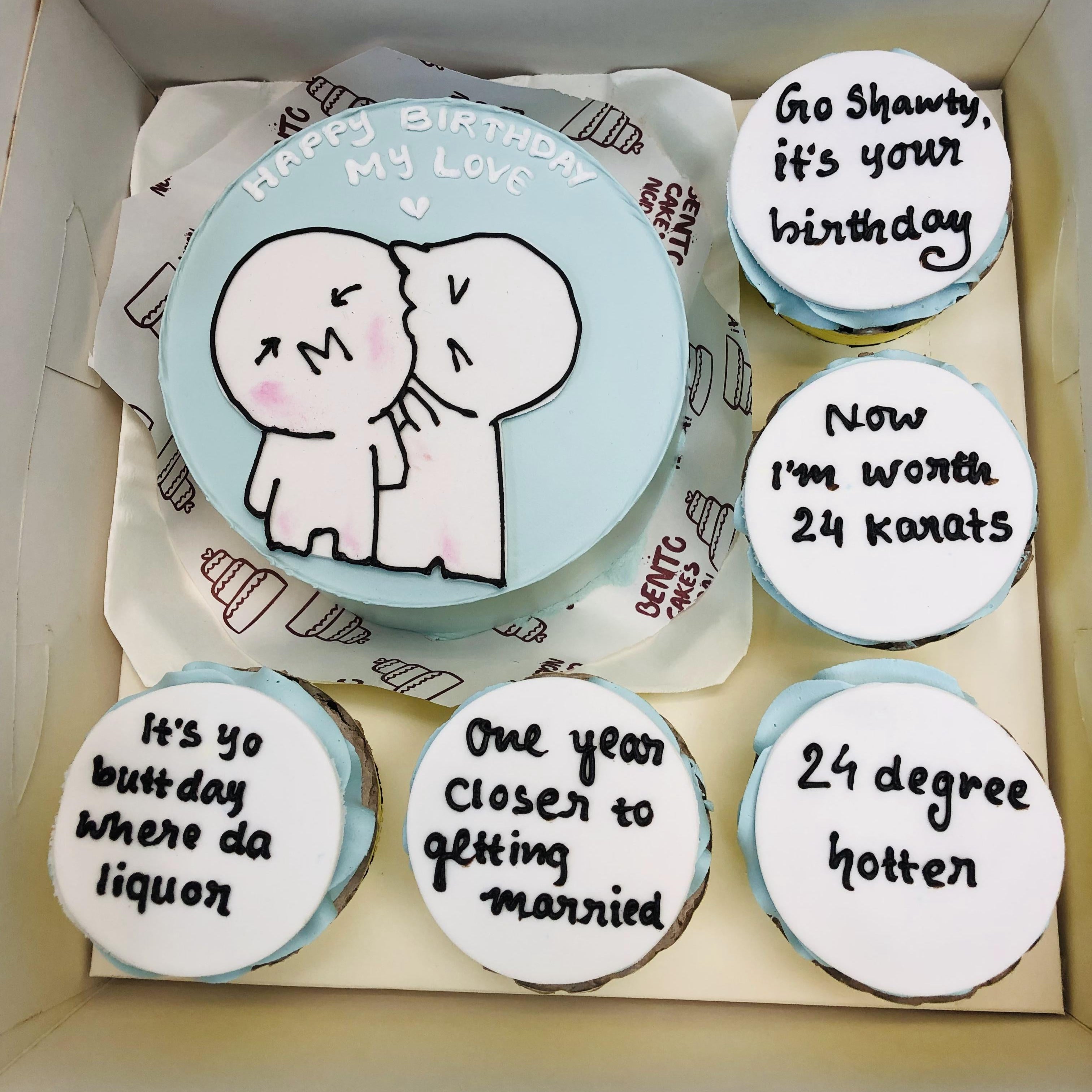 A box containing a light blue frosted birthday cake featuring a cartoon couple kissing and the message "Happy Birthday My Love," alongside five cupcakes, each with personalized playful messages in black icing.