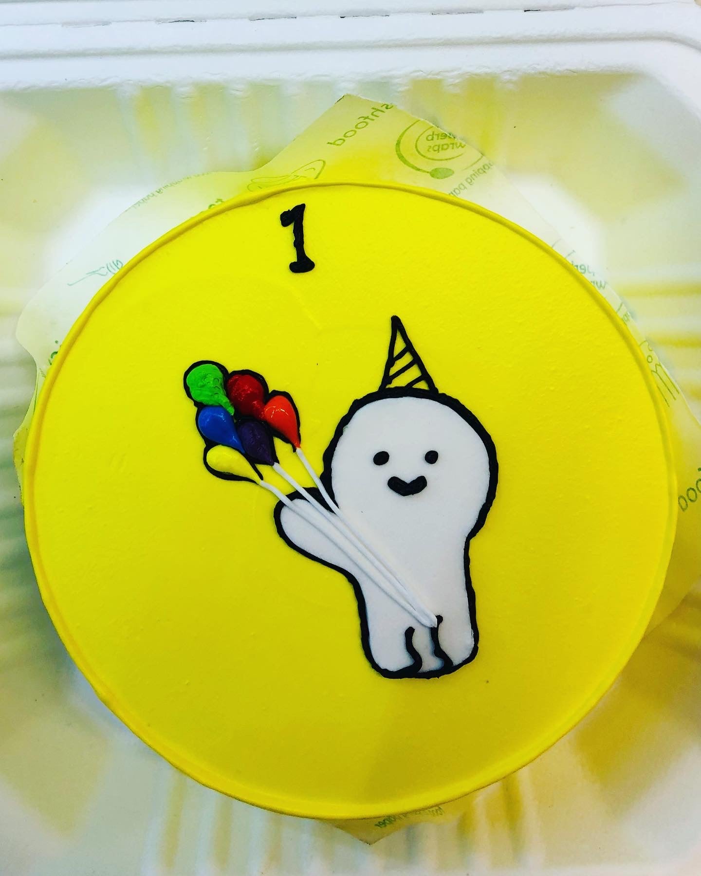 A yellow icing cake is decorated with a cute little cartoon having a bundle of balloons  and a text of 1 written on it. 