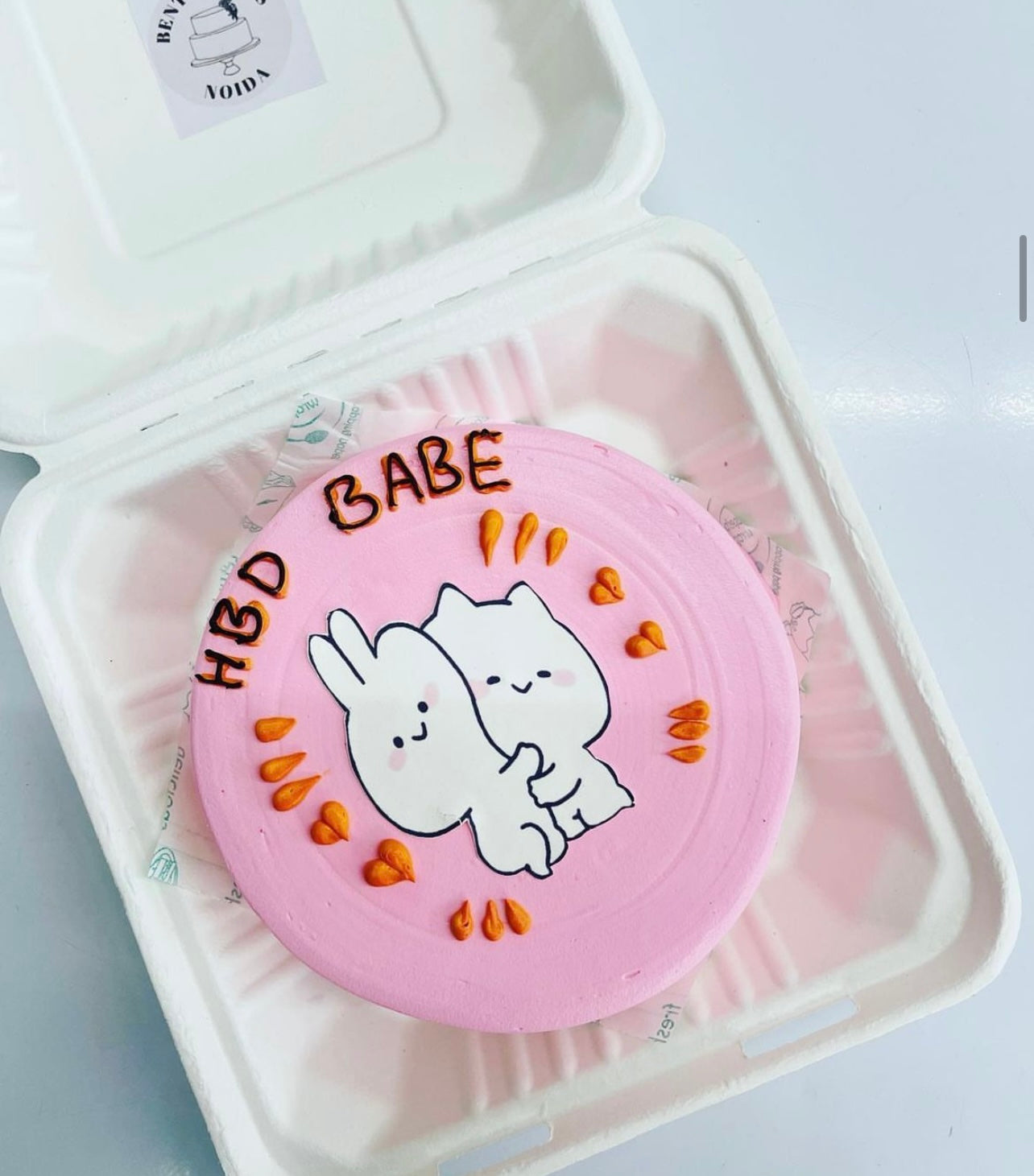 A pink birthday cake with a cute illustration of two hugging characters, a bunny and a bear, surrounded by small orange hearts. The cake is placed inside a bento box. The text "HBD BABE" is written on the cake in black icing.