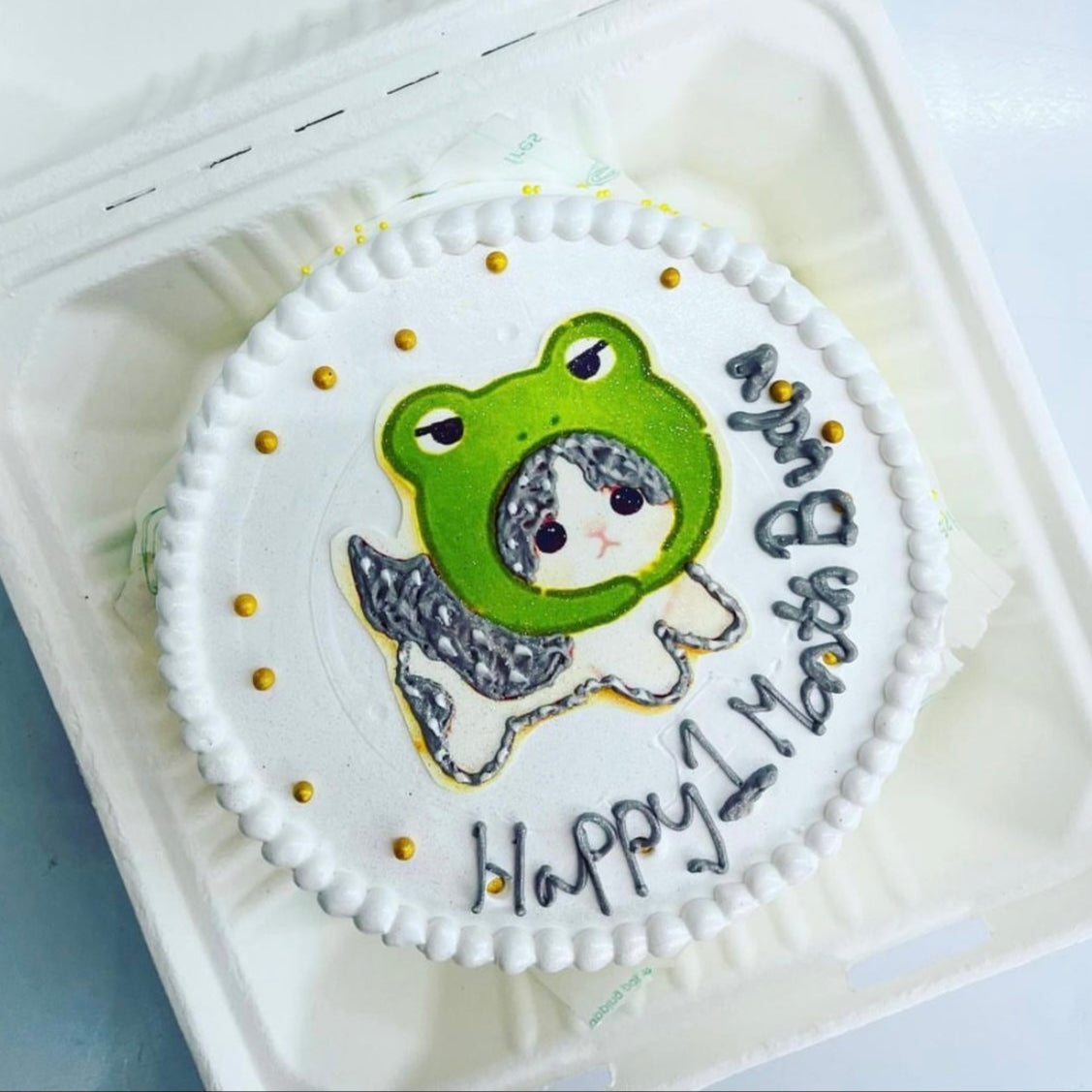 A delightful cake featuring a cute cat wearing frog mask design placed on top, beautifully presented in a charming box.