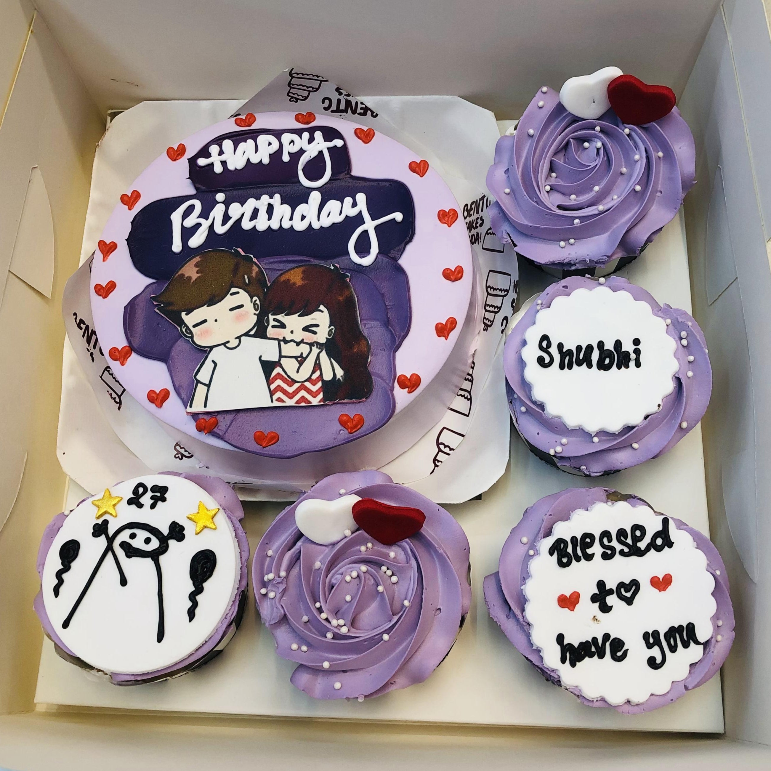 A purple-themed bento cake set featuring a round cake with a drawing of a cute couple and the text "Happy Birthday," surrounded by small red hearts. Accompanying the cake are four cupcakes with personalized designs, including messages like "Shubhi," "Blessed to have you" with red hearts, and a drawing of a stick figure with "27" and two stars. One cupcake has a swirl of purple frosting with heart-shaped decorations.