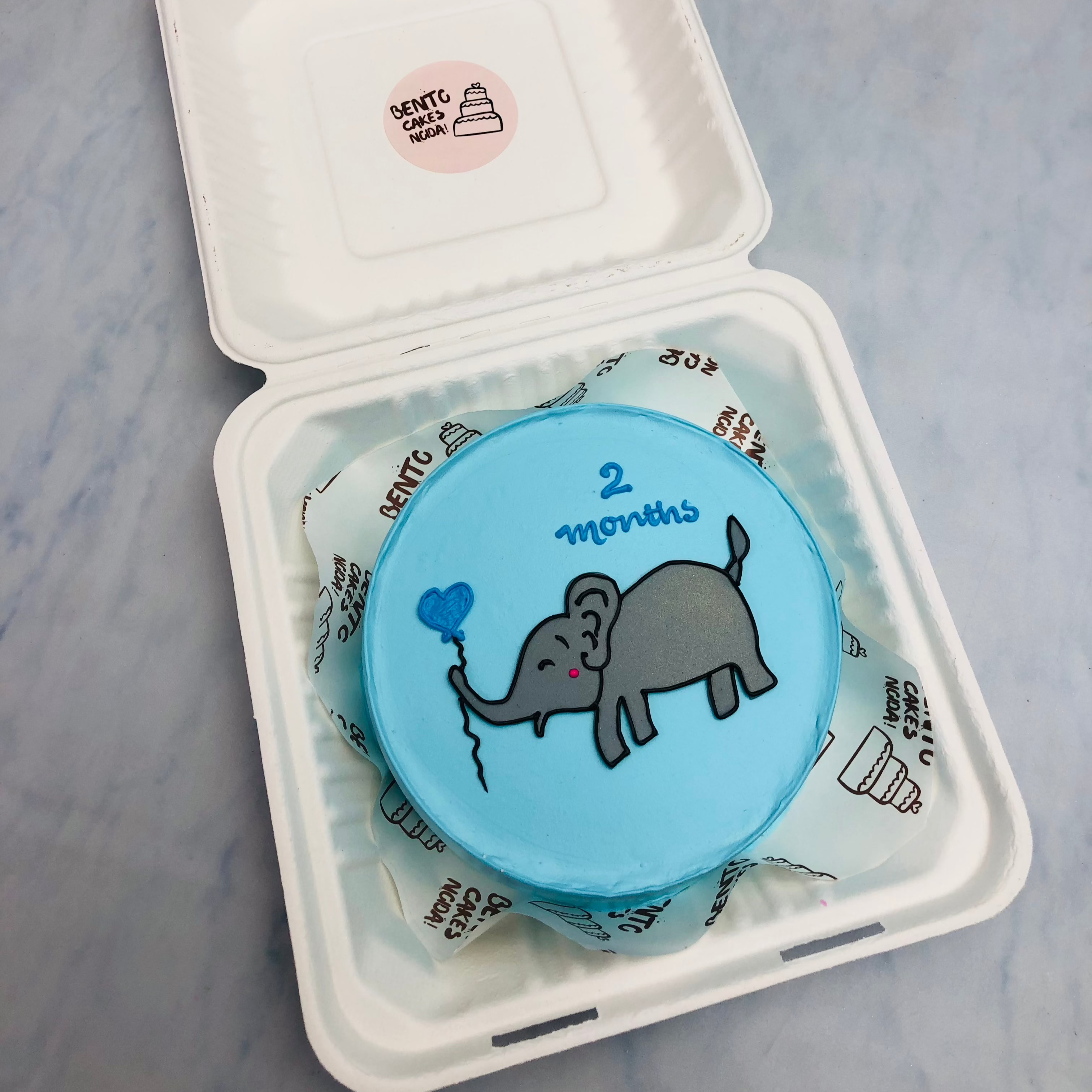 A skyblue bento cake with a cute elephant design on it and taken blue balloon.