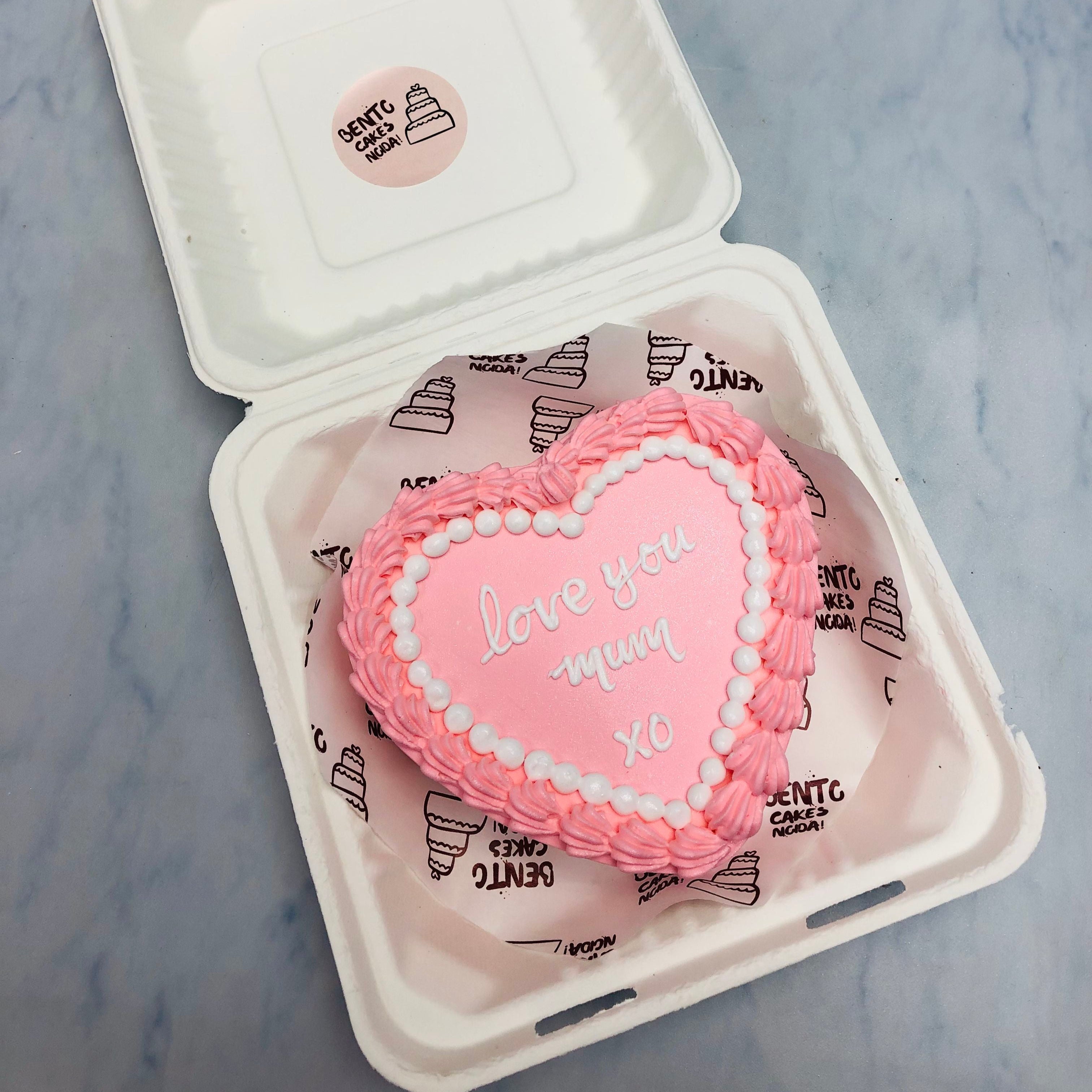 A lighe pink heart shape cake designed for mothers day having taxt "Love Youe Mum XO" on it.