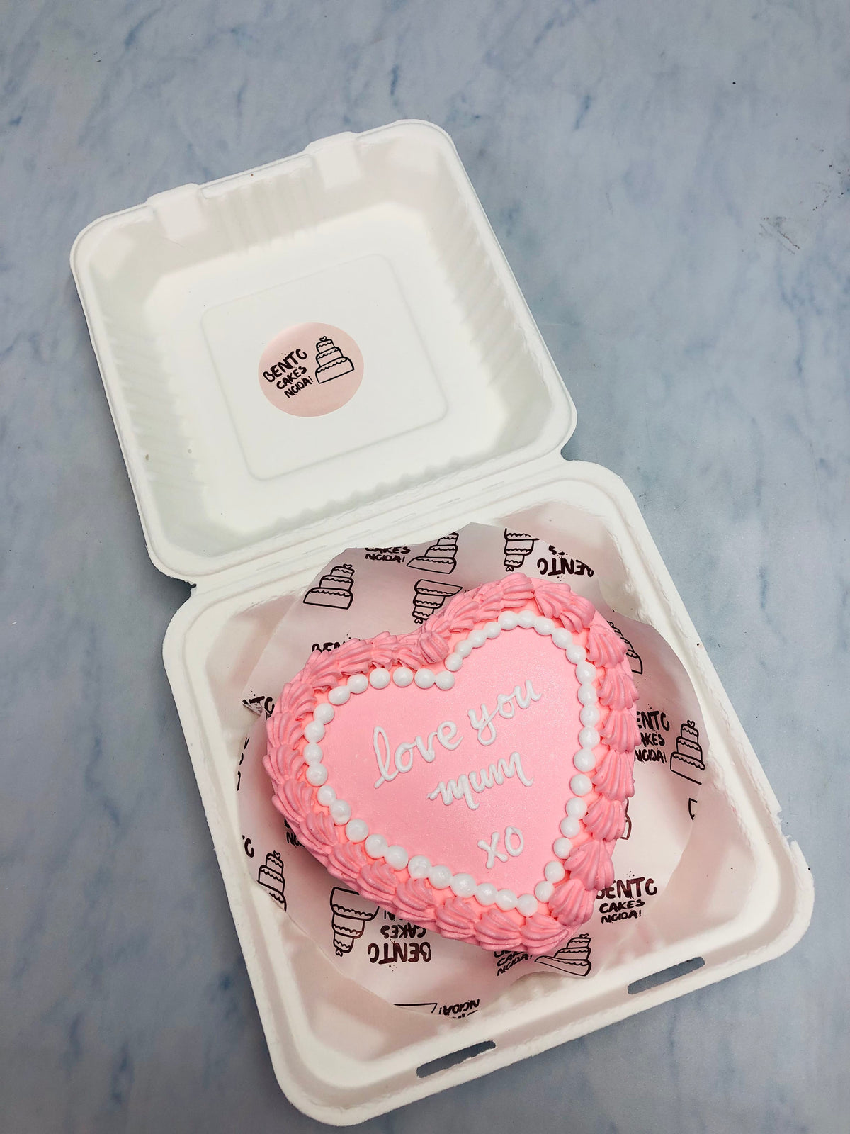 A lighe pink heart shape cake designed for mothers day having taxt "Love Youe Mum XO" on it.