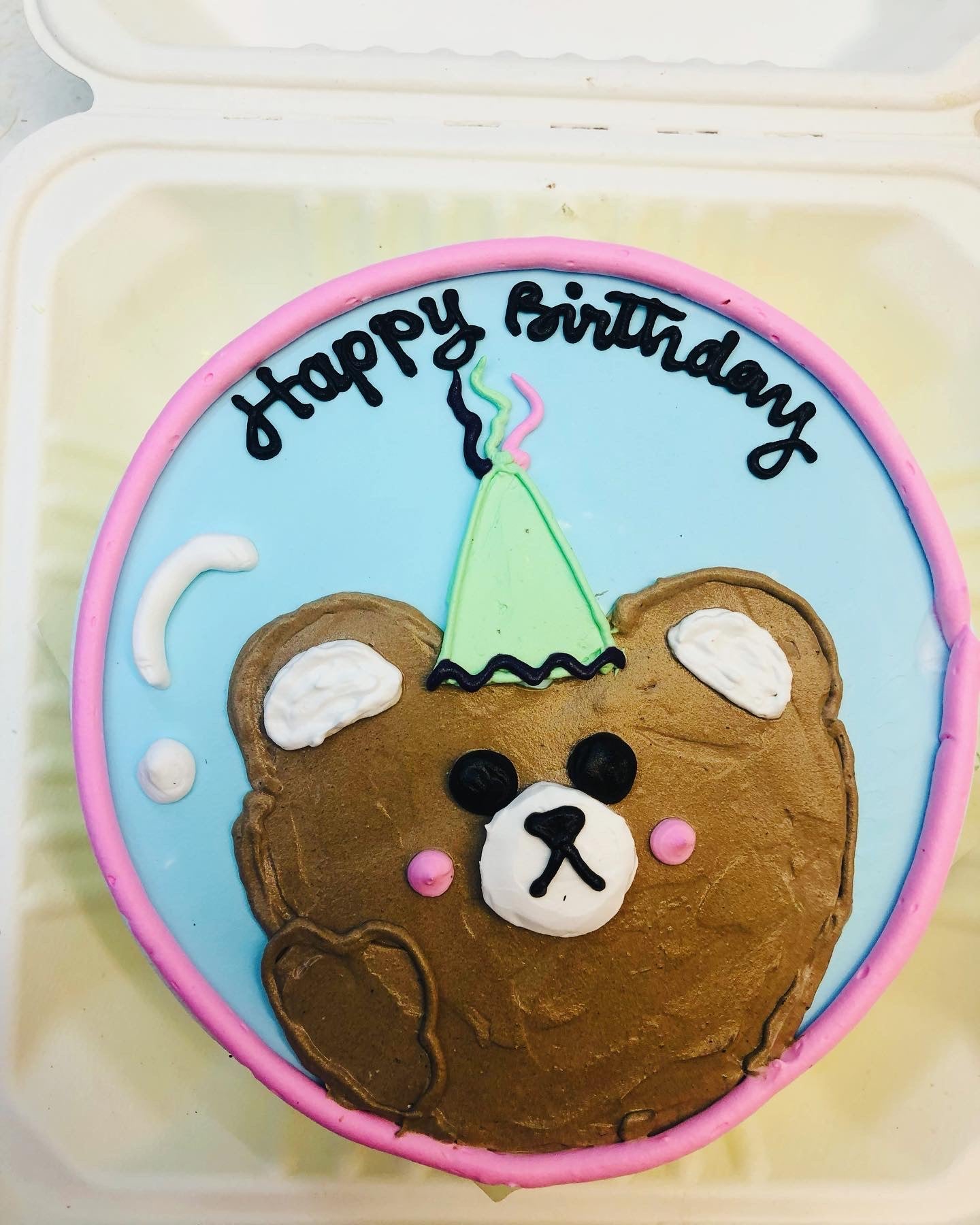 A round birthday cake with a bear face design, wearing a green party hat, and the words "Happy Birthday" written on top.
