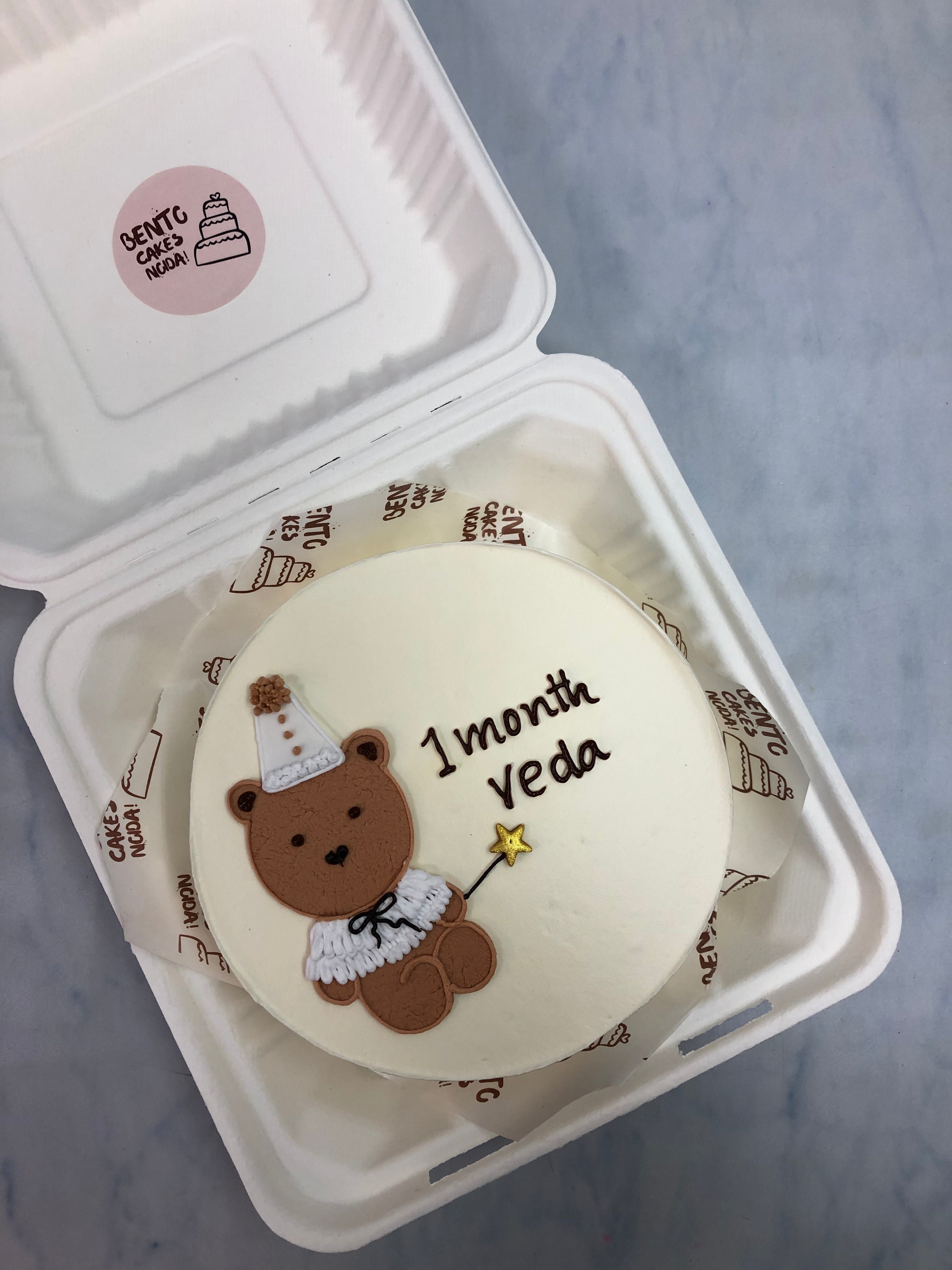A white bento cake with a cute teddy wearing birthday cap and designed for 1 Month baby.