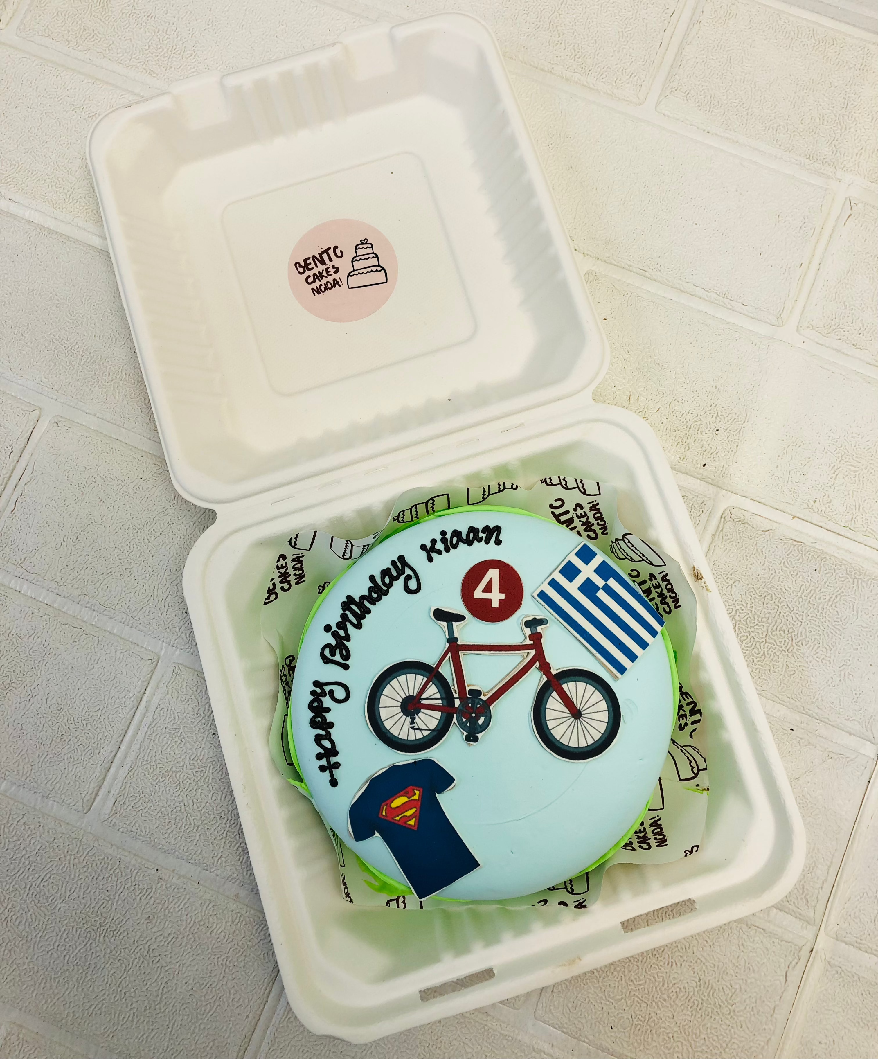 A light blue bento cake having superman tshirt, bicycle and flag theme print.