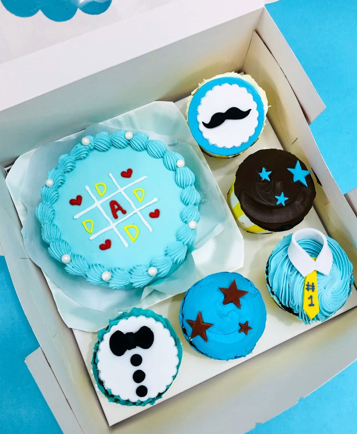 A blue bento cake with Tic Tac Tow design and 5 cupcakes with different designs for dads love.