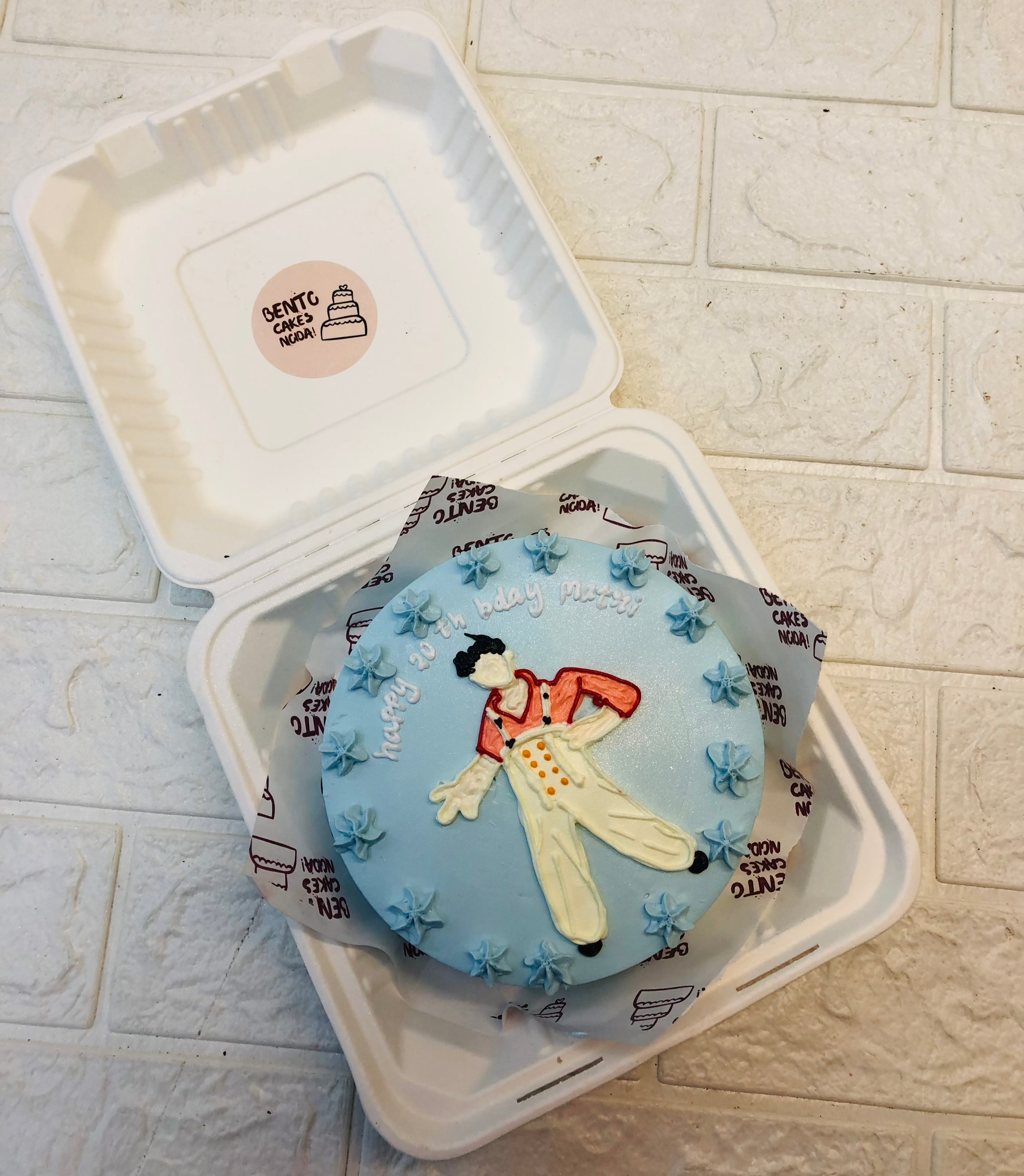 A beautifully decorated light blue bento cake inside a white takeout box, featuring a charming clown design. The clown wears a red and white outfit with yellow pants, surrounded by small blue frosting flowers. The cake has “Happy B-day [name]” written on it, adding a festive touch. 