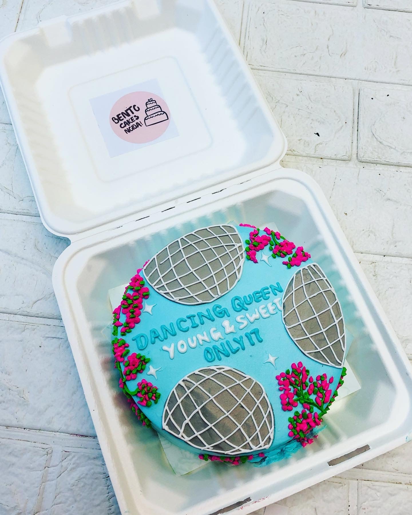 A round cake with a blue frosting base, decorated with four silver disco balls and pink and green floral accents, placed inside a bento box. The cake has the text 'DANCING QUEEN YOUNG & SWEET ONLY 17' written in blue icing.