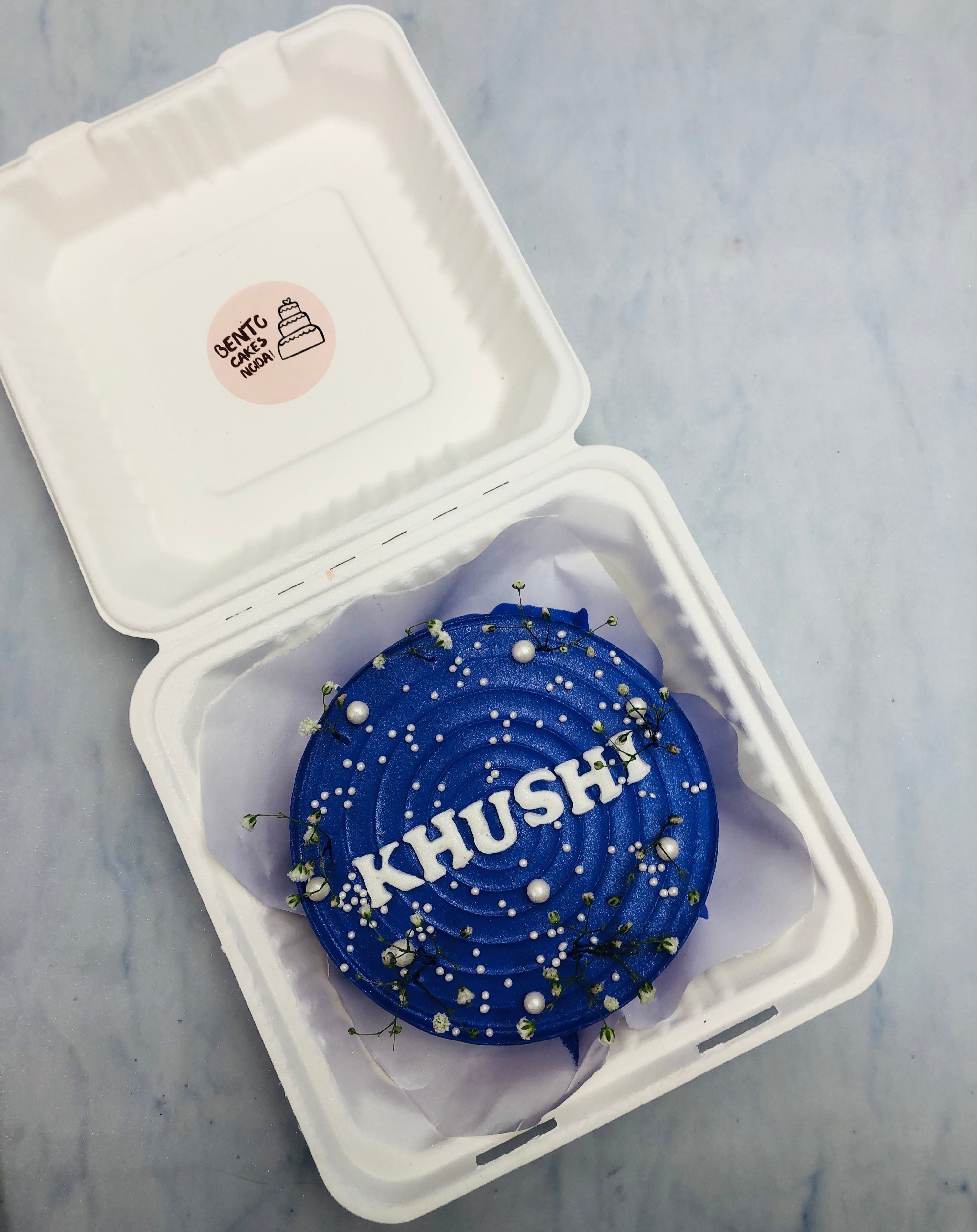 A beautifully crafted blue birthday cake featuring the name "KHUSHI" in white lettering, adorned with small white pearls and delicate green sprigs.
