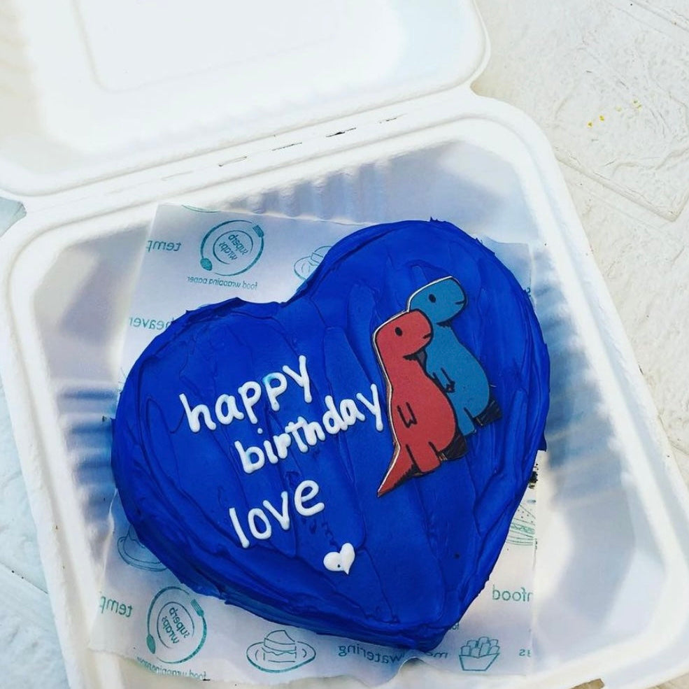 A dark blue heart shape cake is designed for lovers birthday having 2 little dinosaurs print on it and a text "happy birthday love" written with white icing. 