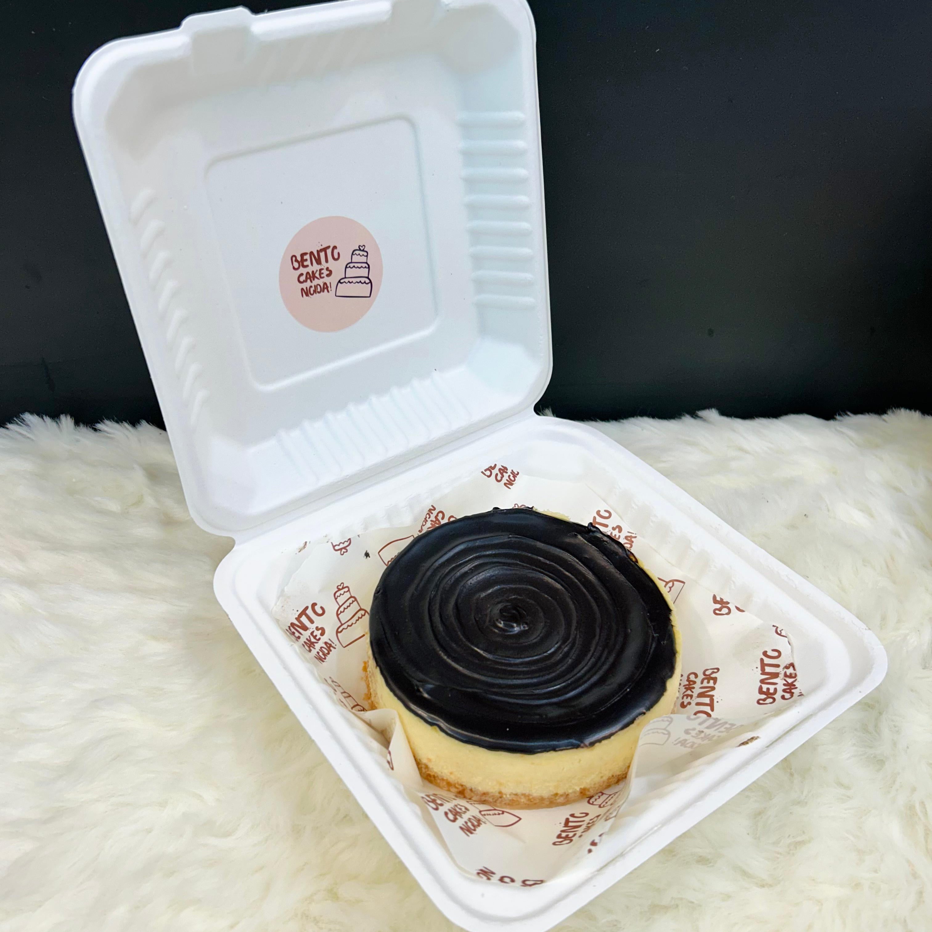 A luxurious Dark Chocolate Cheesecake Bento placed in a white takeaway container lined with branded paper, topped with glossy dark chocolate ganache and garnished with Ferrero Rocher chocolates and chopped nuts, set on a soft white surface.