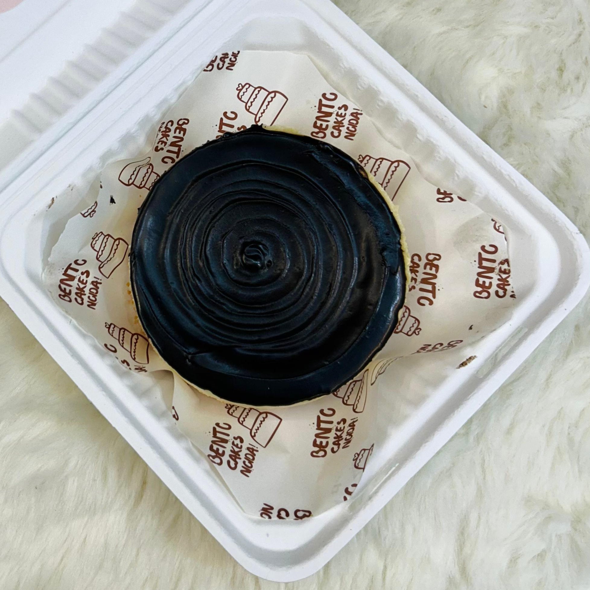A luxurious Dark Chocolate Cheesecake Bento placed in a white takeaway container lined with branded paper, topped with glossy dark chocolate ganache and garnished with Ferrero Rocher chocolates and chopped nuts, set on a soft white surface with front view.