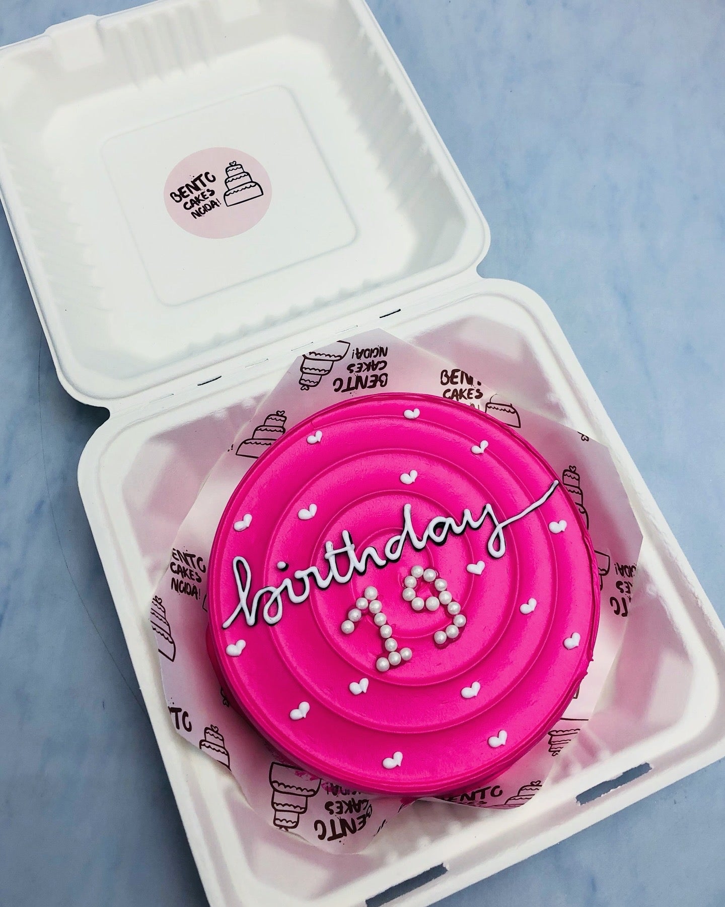 A dark pink frosting bento cake having circular design on top and filled with white hearts and sprinkles and a text "birthday 19" written on it.