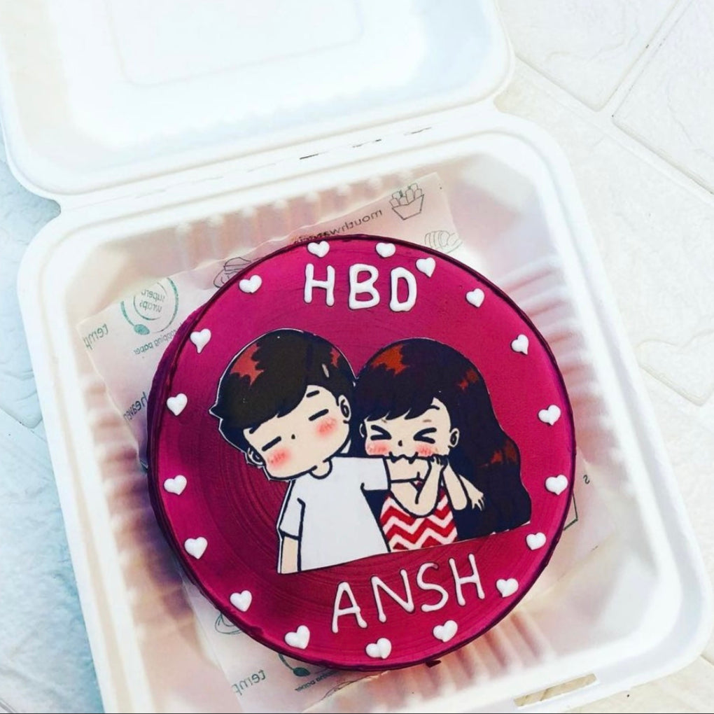 A round birthday cake with a pinkish-red frosting, featuring a cartoon illustration of a boy and a girl. The boy is wearing a white shirt, and the girl is wearing a red and white zigzag patterned dress. The cake has the text "HBD ANSH" written in white icing, surrounded by small white heart decorations. The cake is placed inside a white plastic container.