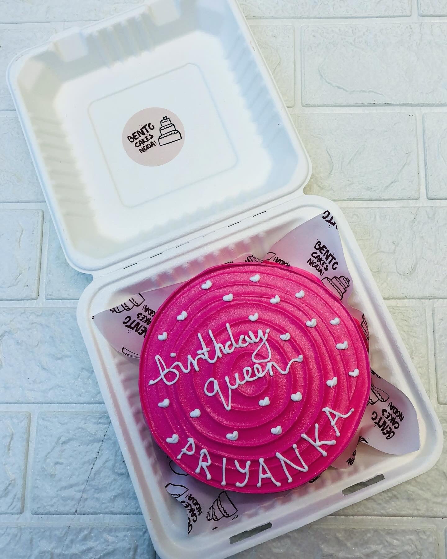 A beautifully crafted pink birthday cake adorned with white icing that spells out 'Birthday Queen' and 'PRIYANKA.' The cake is embellished with small white heart decorations.
