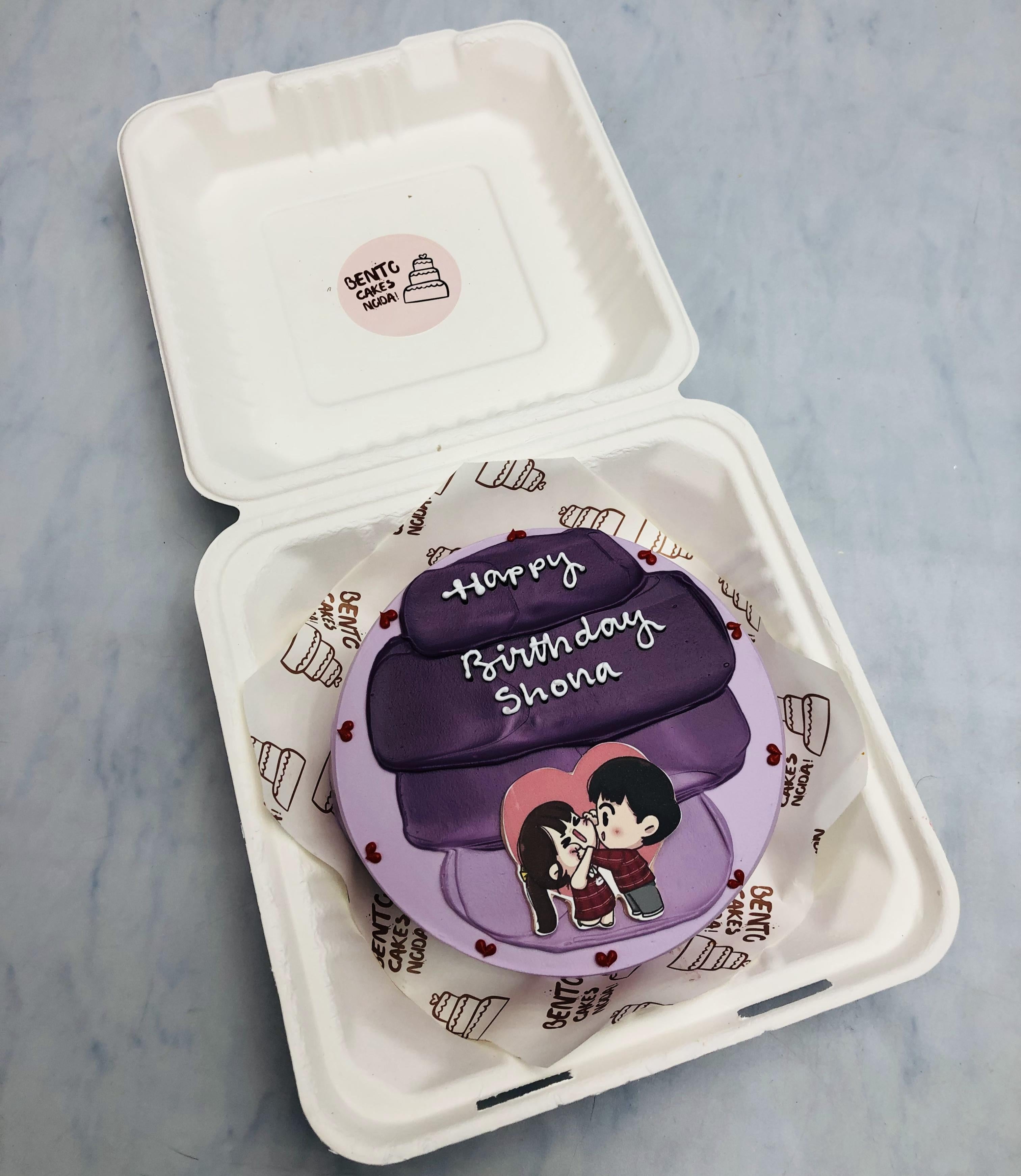 A purple birthday cake with the text "Happy Birthday Shona" and a cute illustration of a couple in a loving pose. 